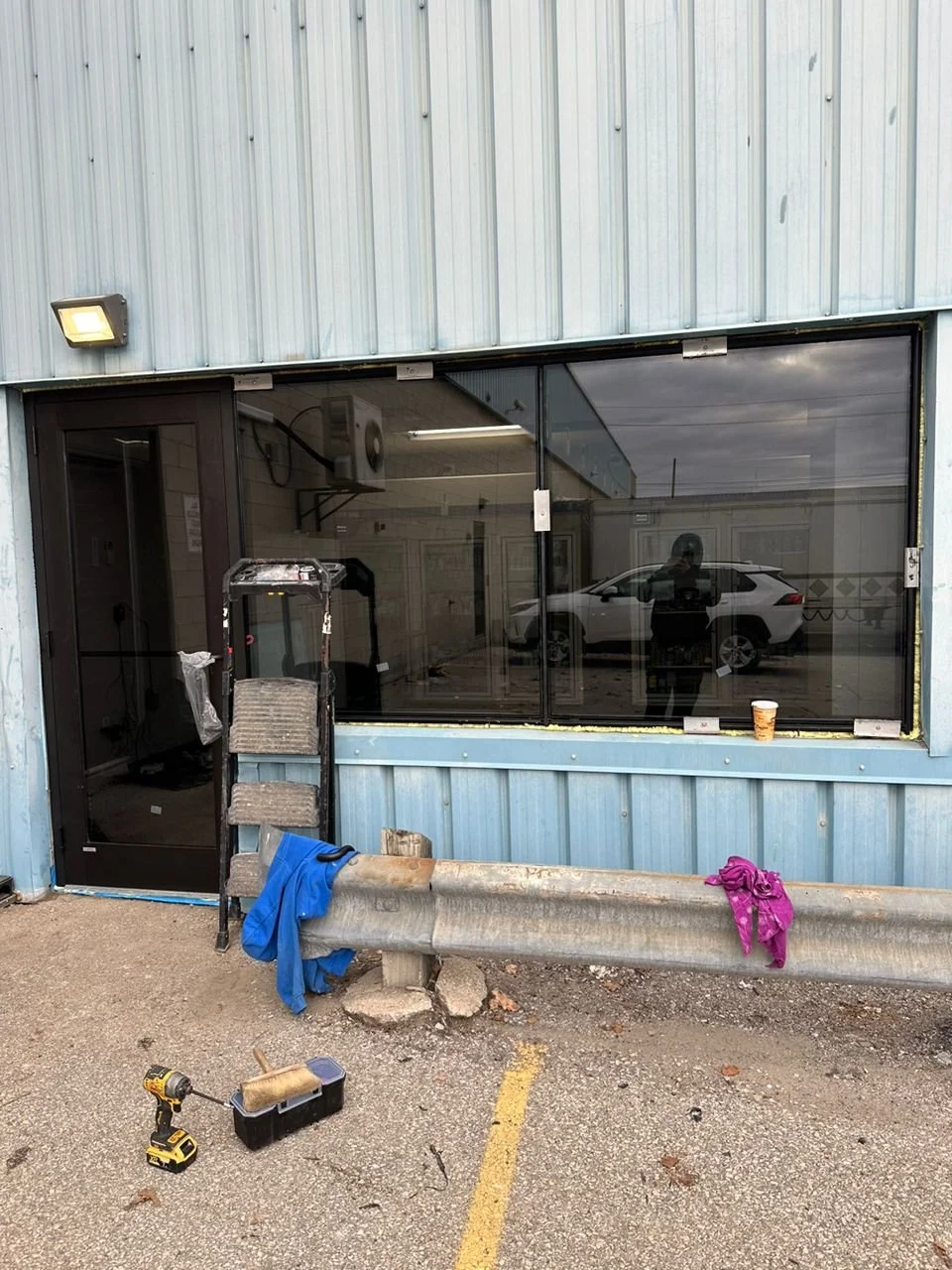 Construction site outside a blue building with a glass window, a step ladder, a yellow drill, a toolbox, and clothing hanging on a metal barrier.