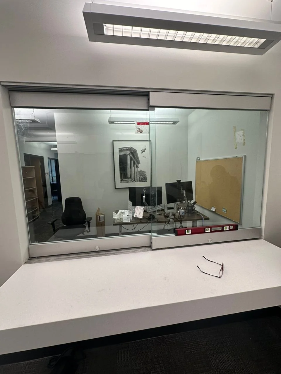 Office reception window with a white countertop, interior view of an office with a chair, computer monitors, a bulletin board, and framed artwork, viewed through the glass window from outside.