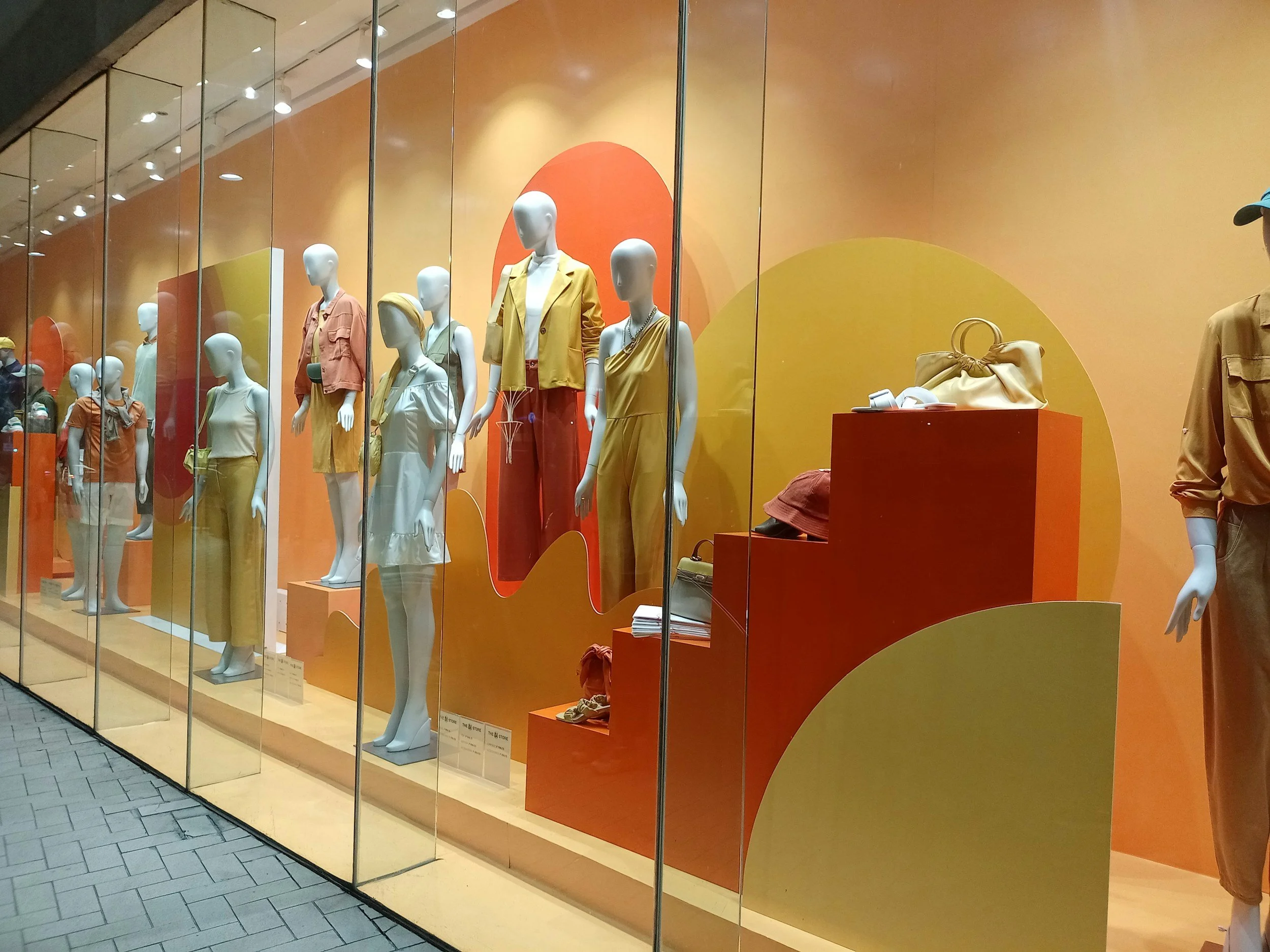 Clothing store display window with mannequins dressed in casual summer outfits in shades of orange, yellow, and beige, with abstract orange and yellow background.