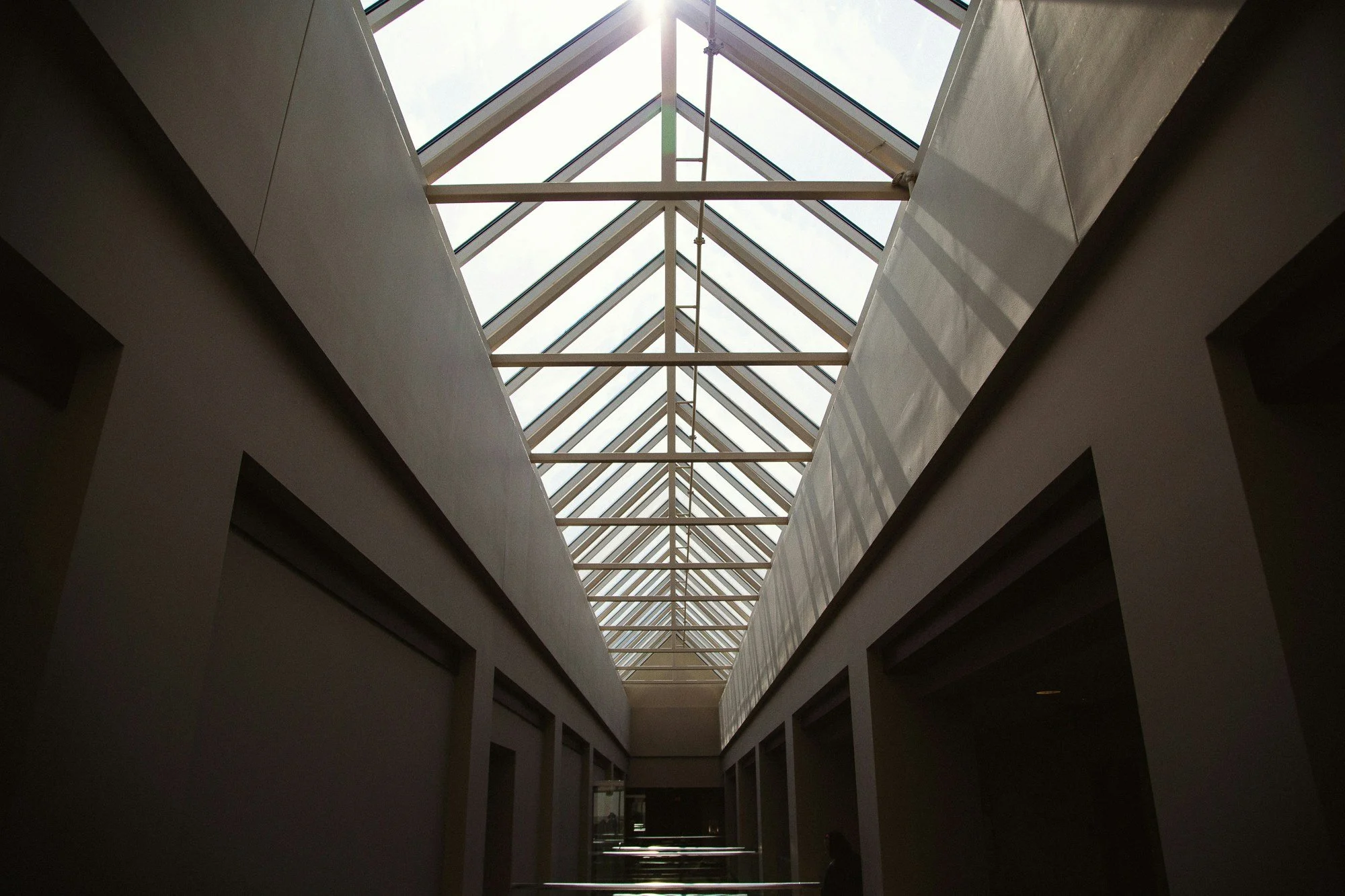 Interior view of a building with a glass ceiling and metal framework, showing a narrow corridor with high walls.