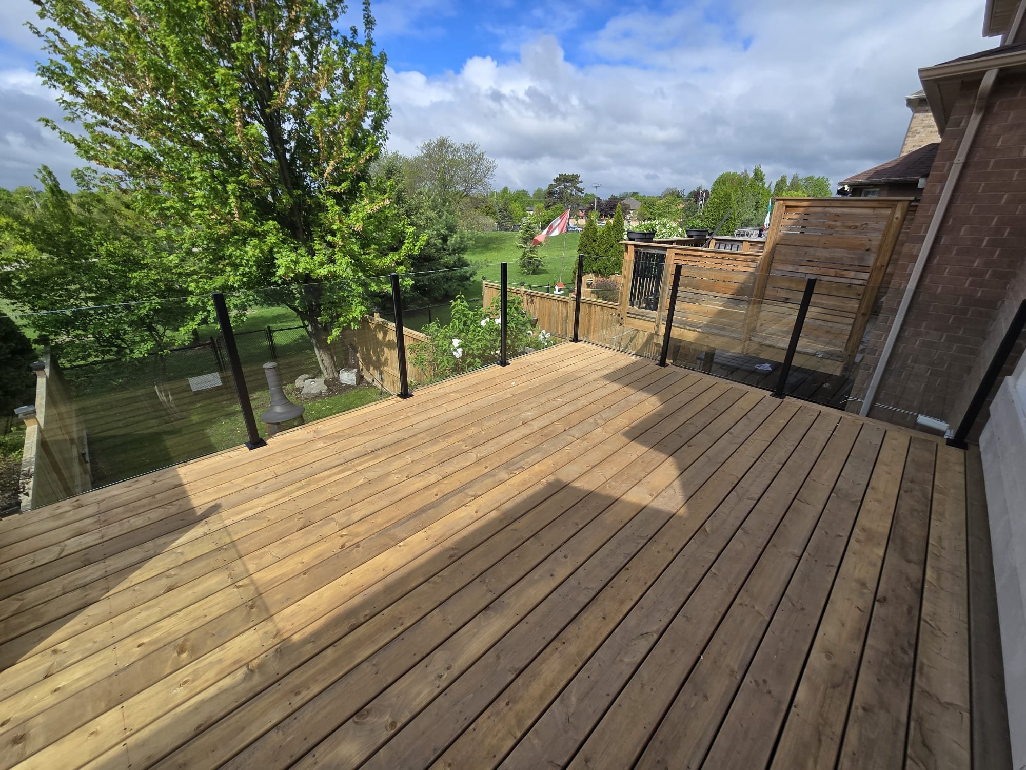 Newly constructed wooden deck with black metal and glass railing overlooking a green backyard with trees and a lawn in a suburban neighborhood.