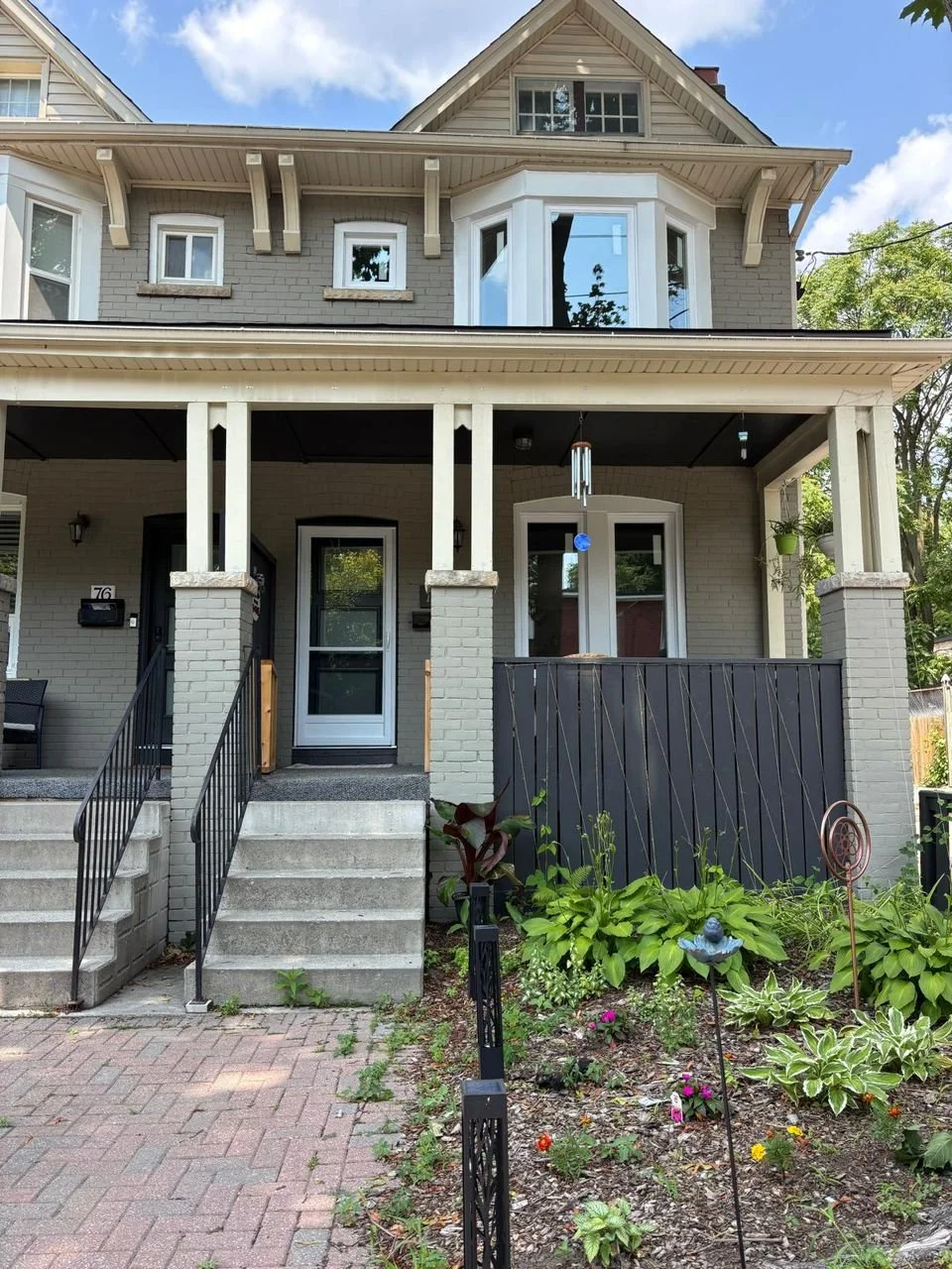 Front of a multi-story house with a porch, stairs, and a garden, featuring gray brick walls, white trim, and a wind chime hanging near the entrance.