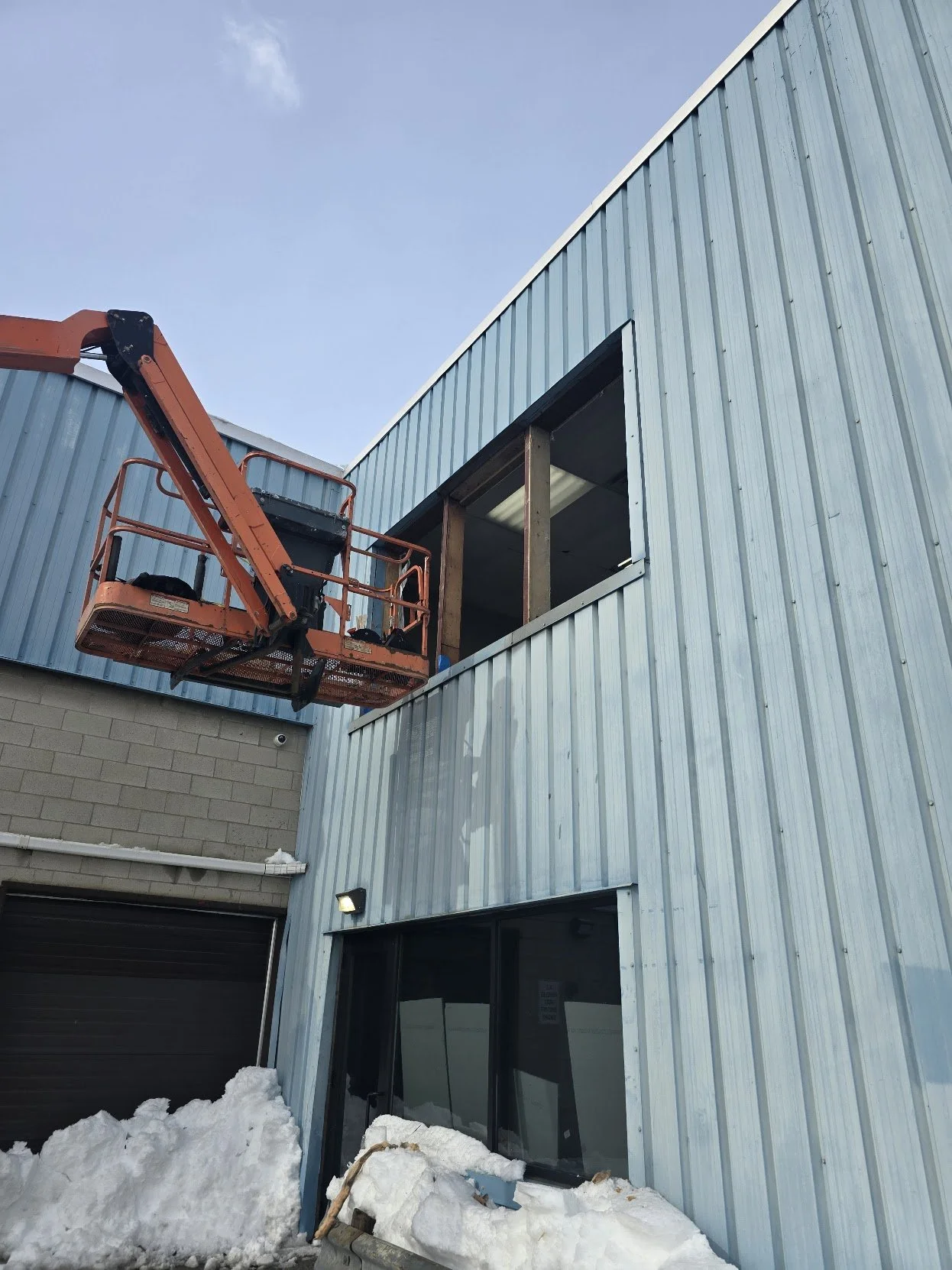 Construction site with building in progress, blue metal siding, window, snow on ground, and an orange lift lift platform.