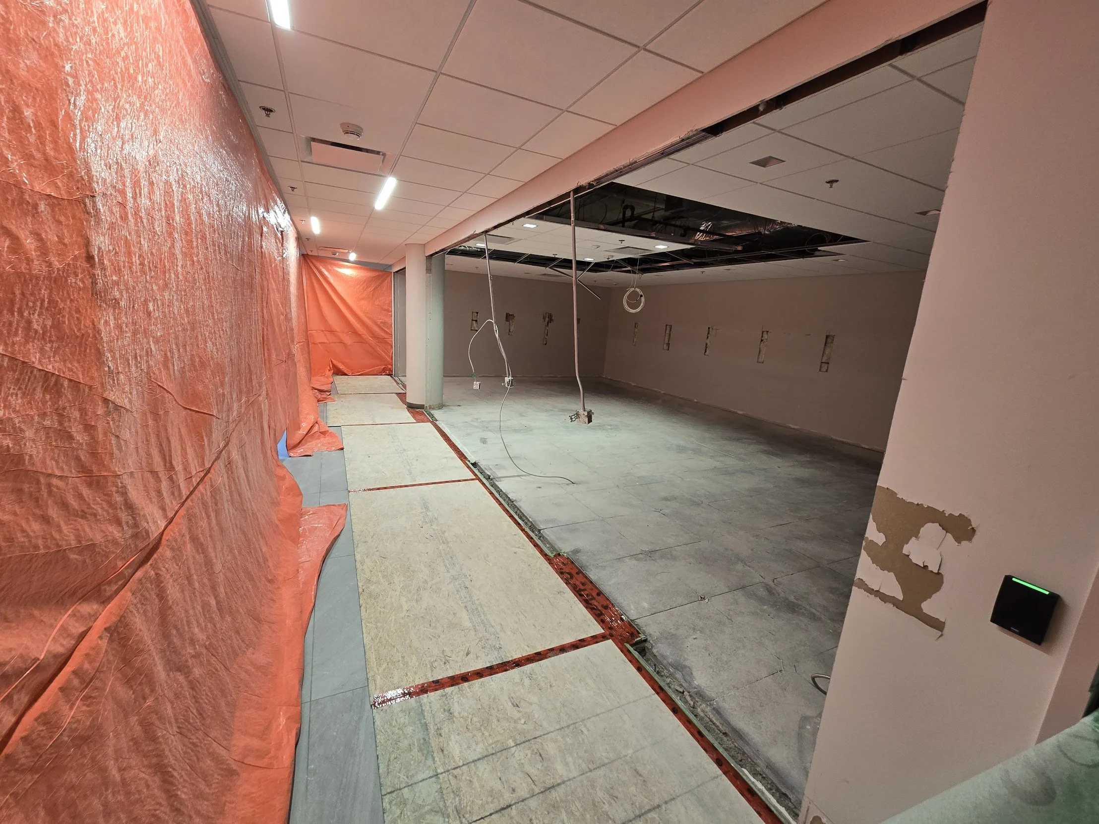 Indoor construction site with partially removed flooring, exposed walls, and ceiling panels. Orange plastic sheeting covers part of the wall, and construction wires hang from the ceiling.