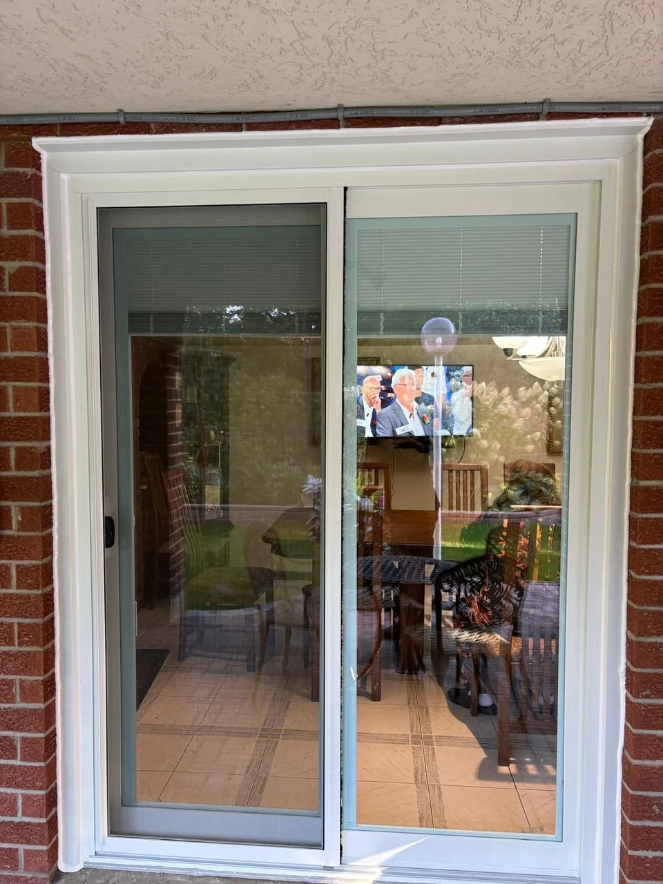 A sliding glass door with a view into a dining area. The reflection shows an outdoor scene with trees and a person taking the photo. Inside, there are chairs, a table, and a television mounted on the wall.