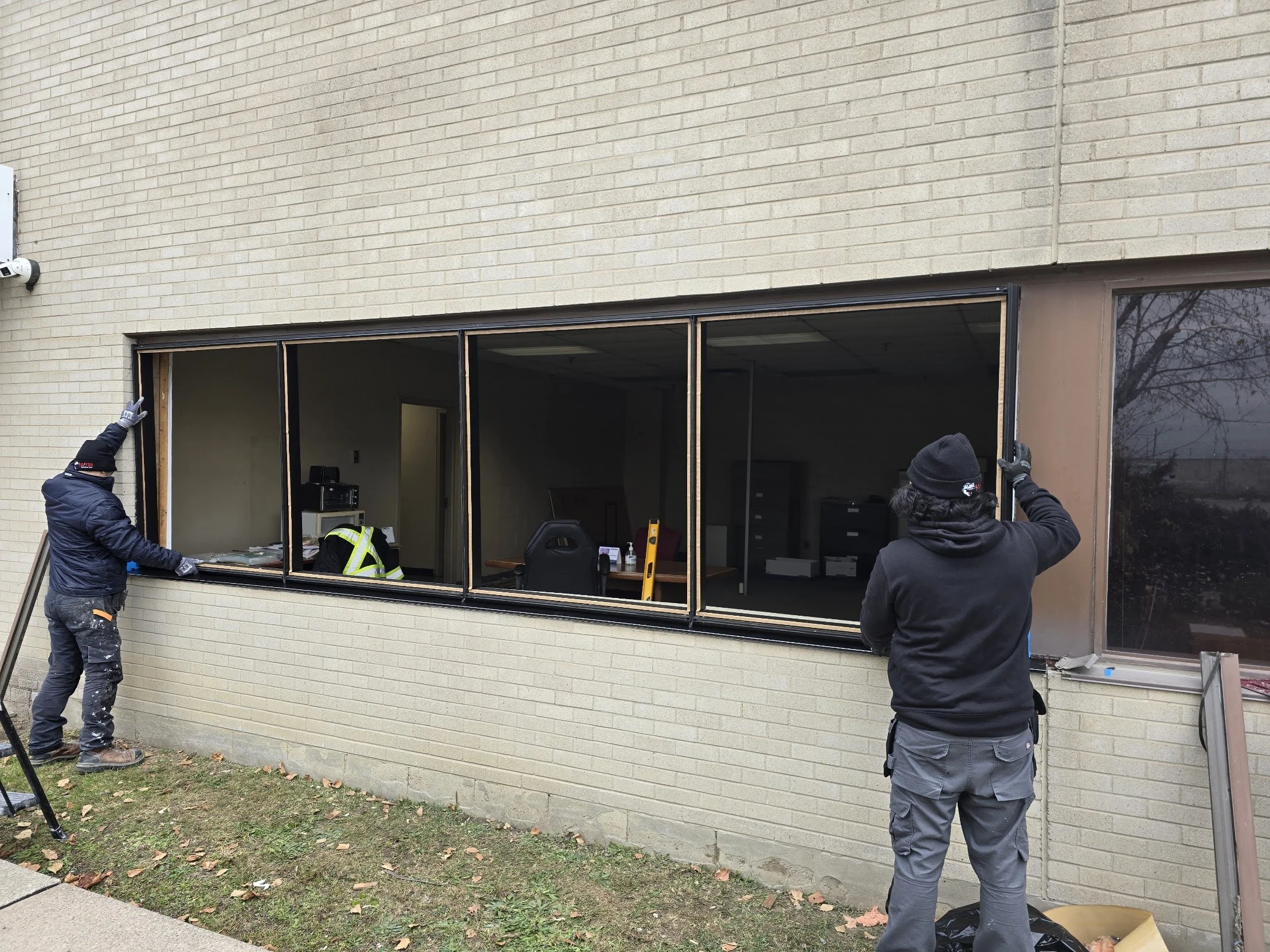 Two workers installing a large window in a brick building. One worker is on the left, holding the window frame, while the other is on the right, pushing the frame into place. Inside the building, visible through the window opening, there is office furniture and equipment.