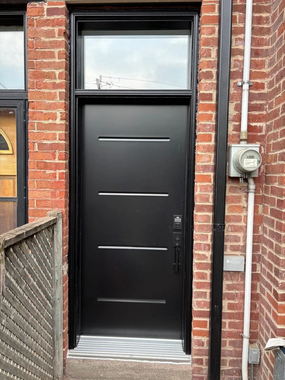 Black modern front door with horizontal silver lines, digital keypad lock, and handle, set in a red brick wall with a window above, next to an electrical meter and conduit.