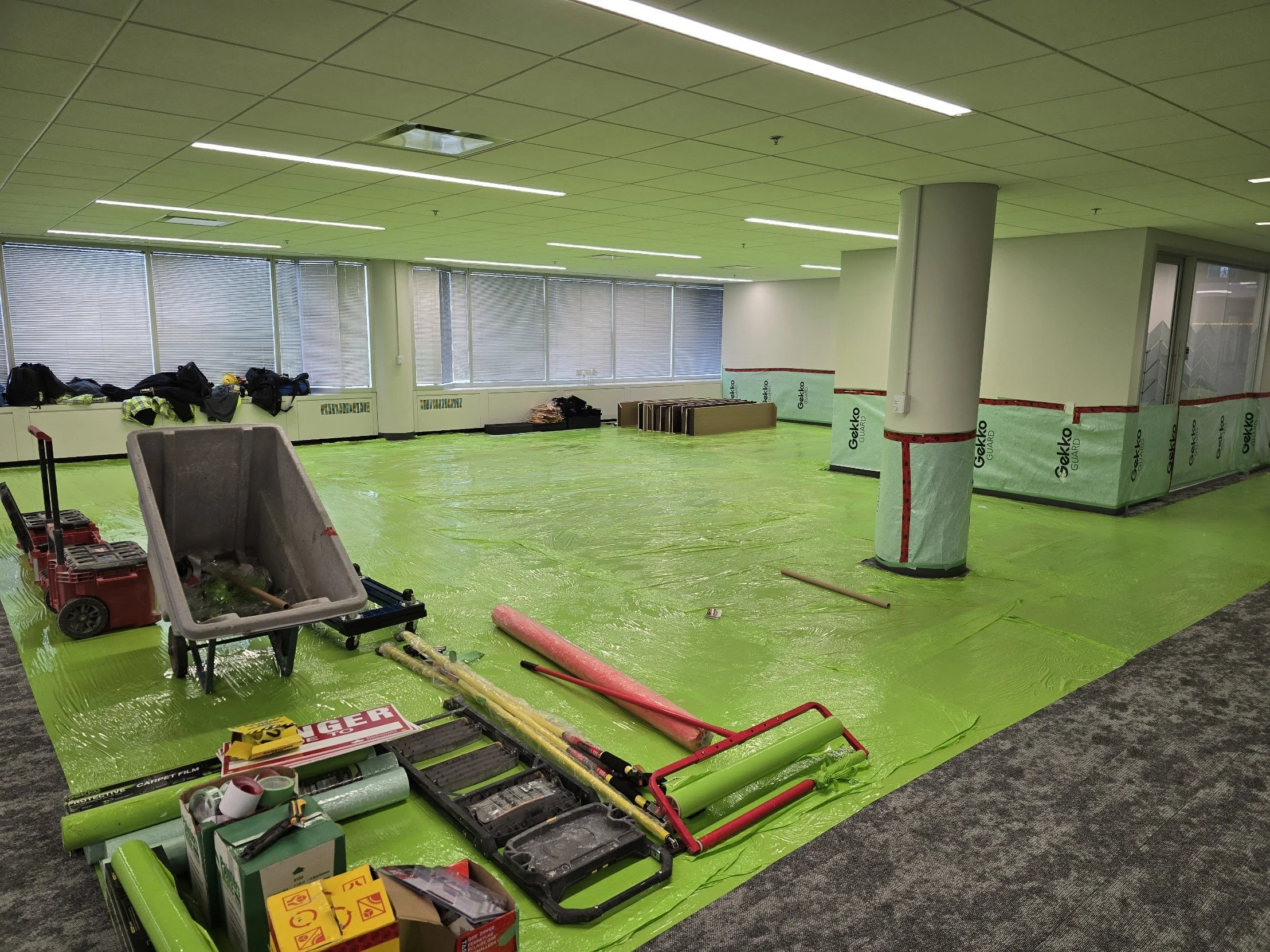 An office under renovation with green protective floor covering, construction tools, and materials spread across the room, with windows and a white ceiling.