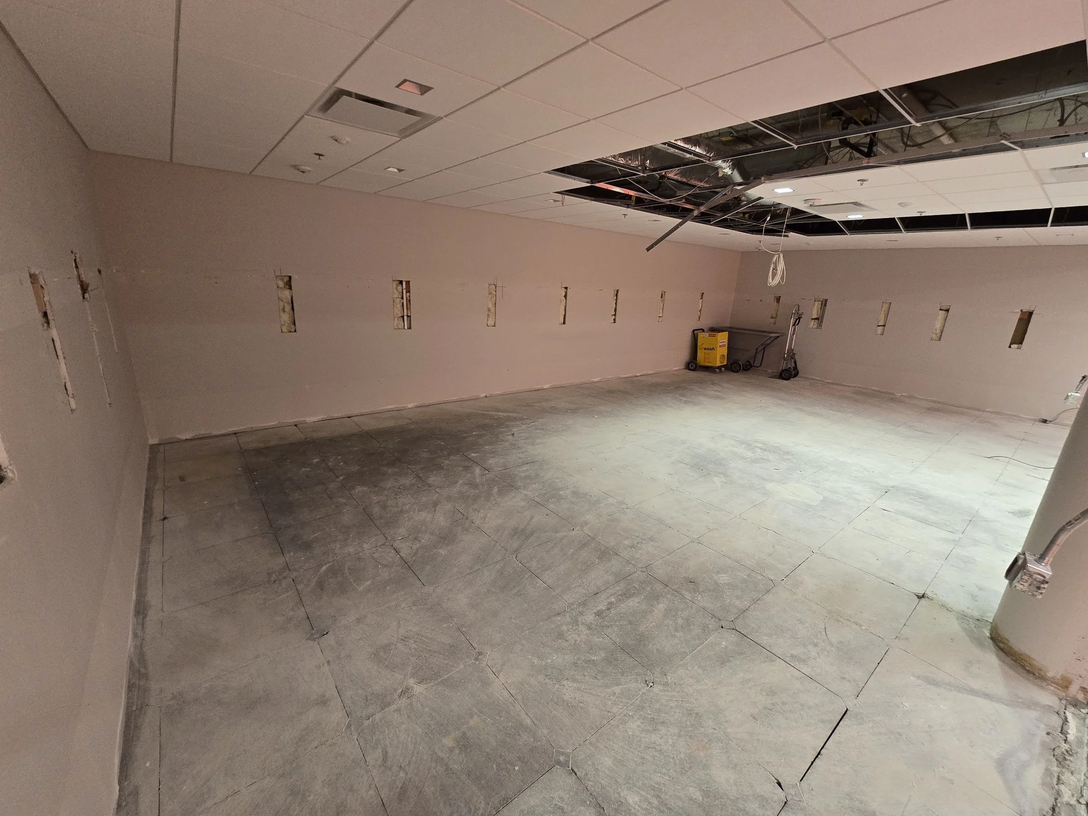 An unfinished room with drywall partially installed on the walls and ceiling, exposed ceiling tiles, and a concrete floor under construction, with construction tools in the corner.