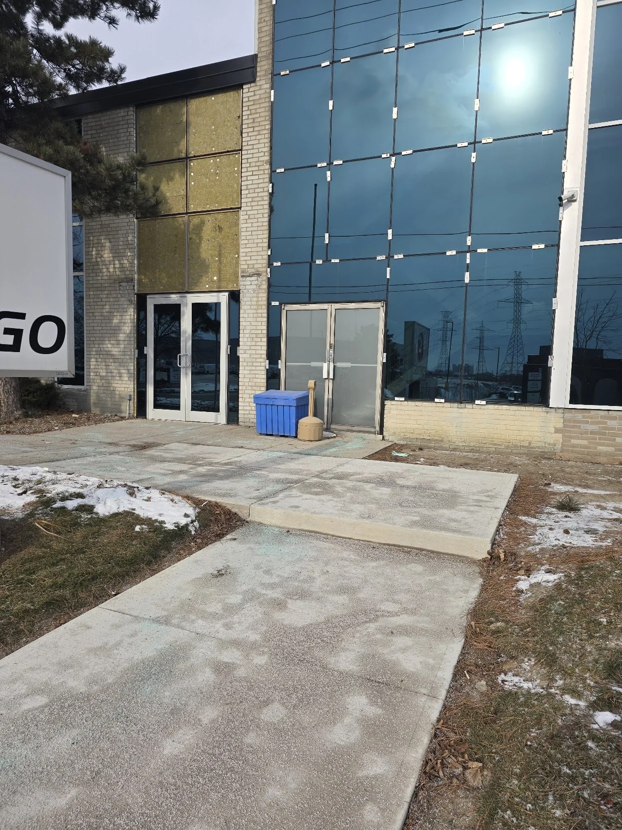 Side view of a commercial building under construction or renovation with reflective blue glass windows, a glass door, a blue recycling bin, and a sidewalk with patches of snow and grass.