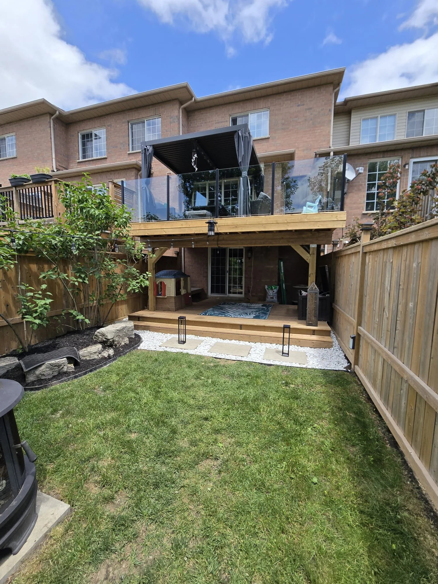 Backyard with a grassy lawn, a wooden deck with steps, a fenced patio area with string lights, and a backyard porch with a screened banister and outdoor furniture under a blue sky with clouds.