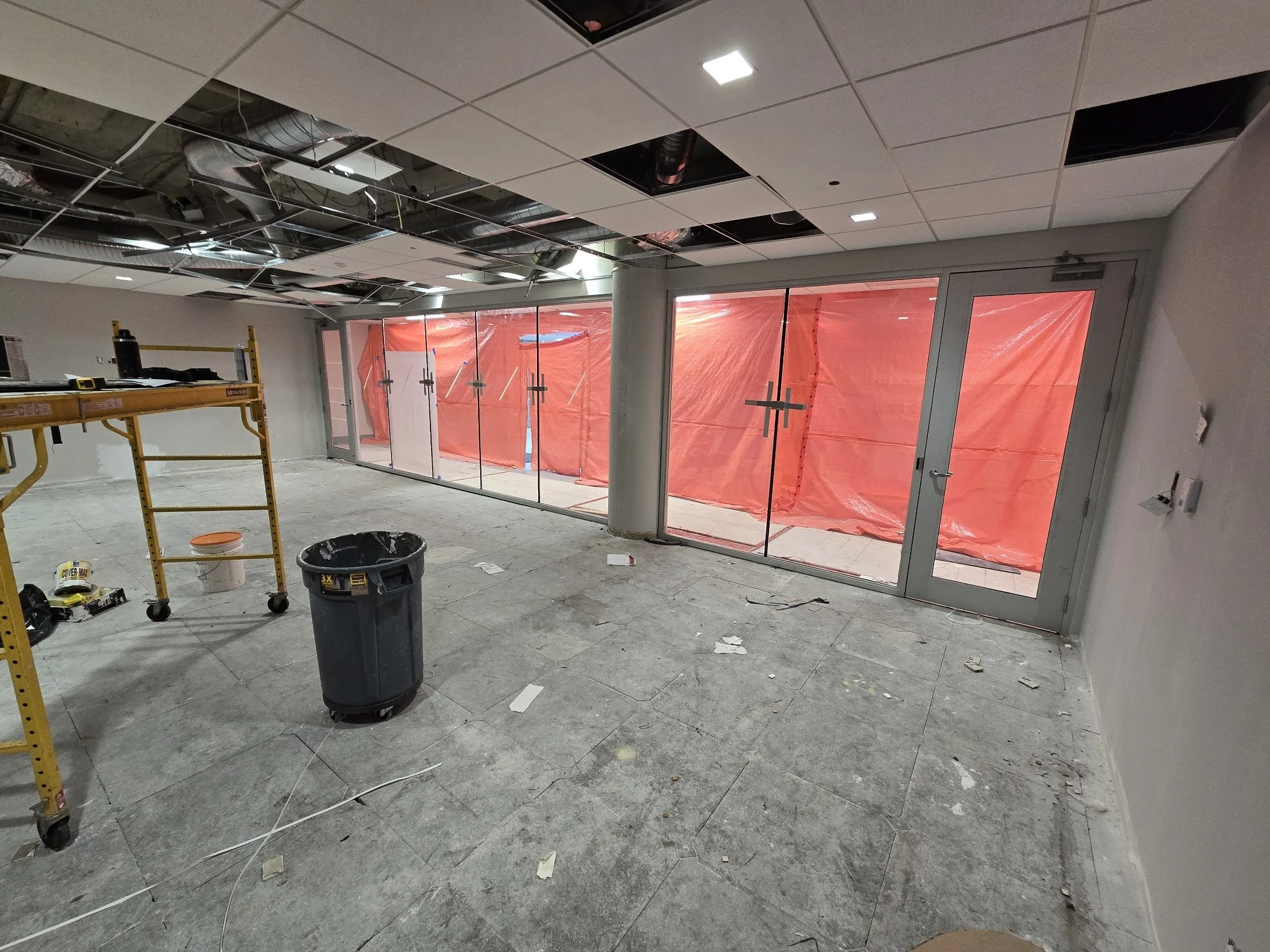 Interior of a room under construction with a scaffold, trash bin, and debris on the floor, and large glass doors covered with red plastic sheeting.