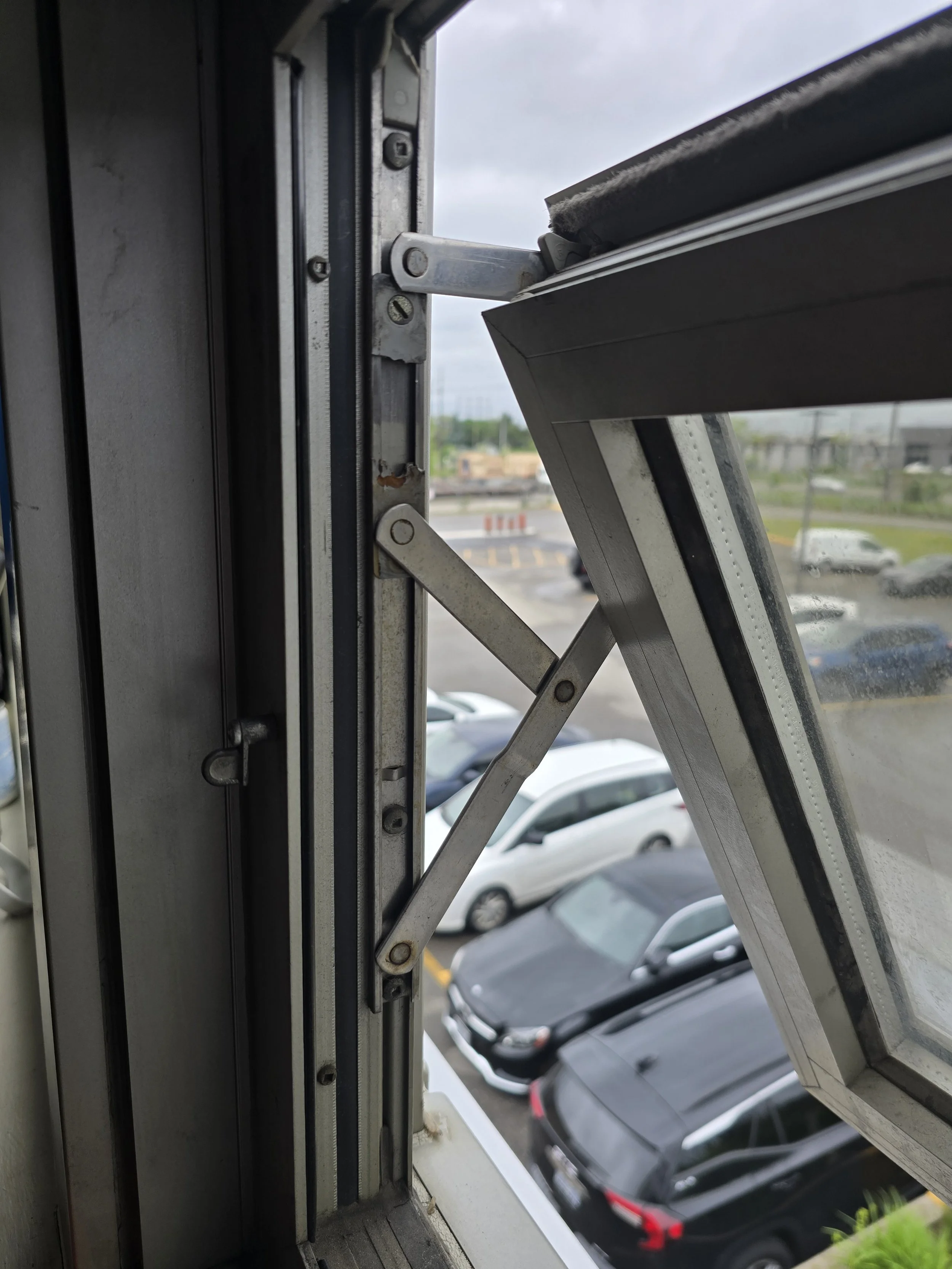 Open window with metal hinges and scissor arm support, overlooking a parking lot with cars and cloudy sky.