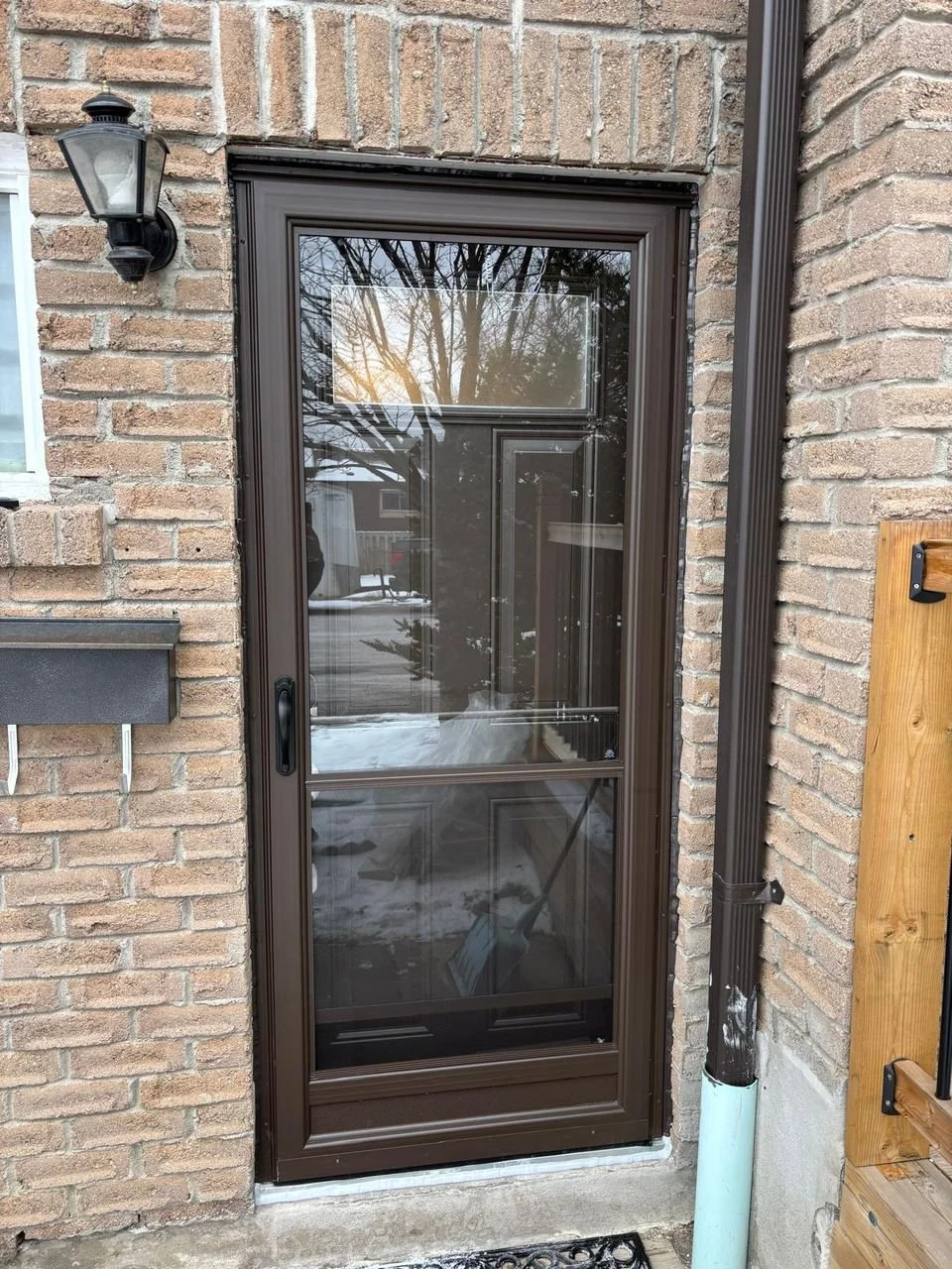 A brown metal screen door with a glass panel is installed in a brick wall outside a house. There is a black outdoor wall lantern light fixture on the left side of the door and a black drain pipe on the right side. A wooden board with black metal brac
