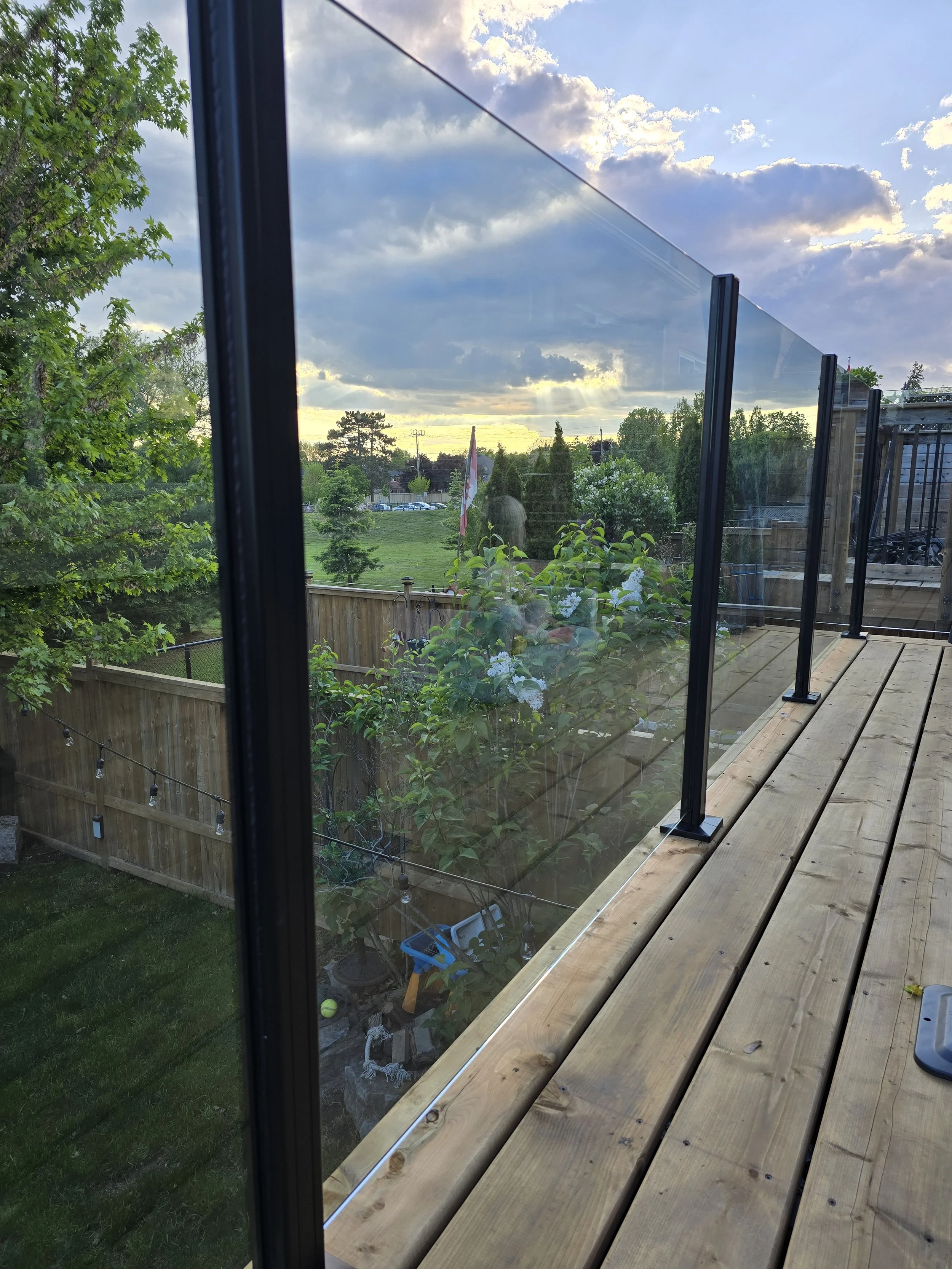 View through a glass railing on a wooden deck overlooking a backyard with trees, a flag, and a sky with clouds at sunset.
