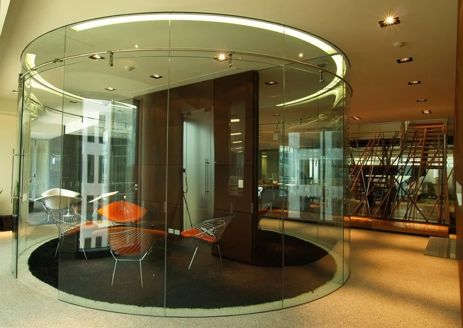 Modern office interior with a glass-walled meeting room, metal chairs with orange cushions, a black rug, and a staircase with wooden steps and metal railings in the background.