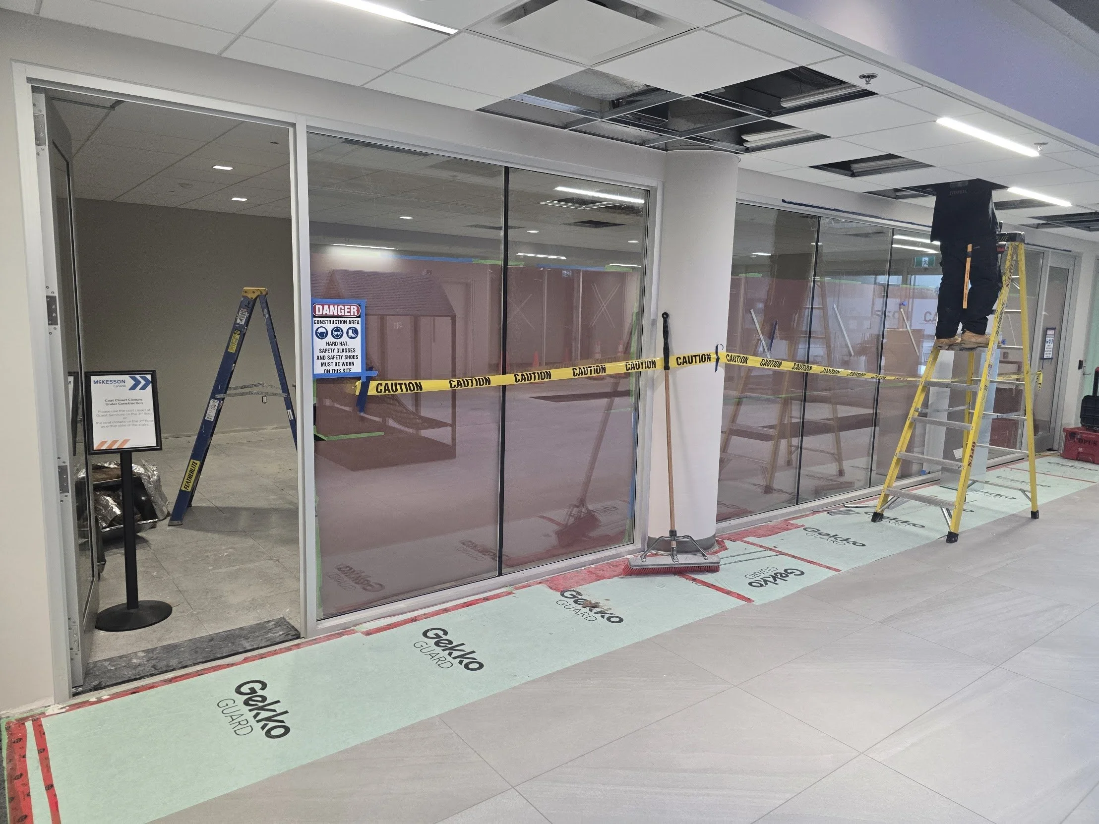 Construction workers installing glass at an indoor storefront. Yellow caution tape cordons off the area, and there are ladders, construction tools, and warning signs visible.