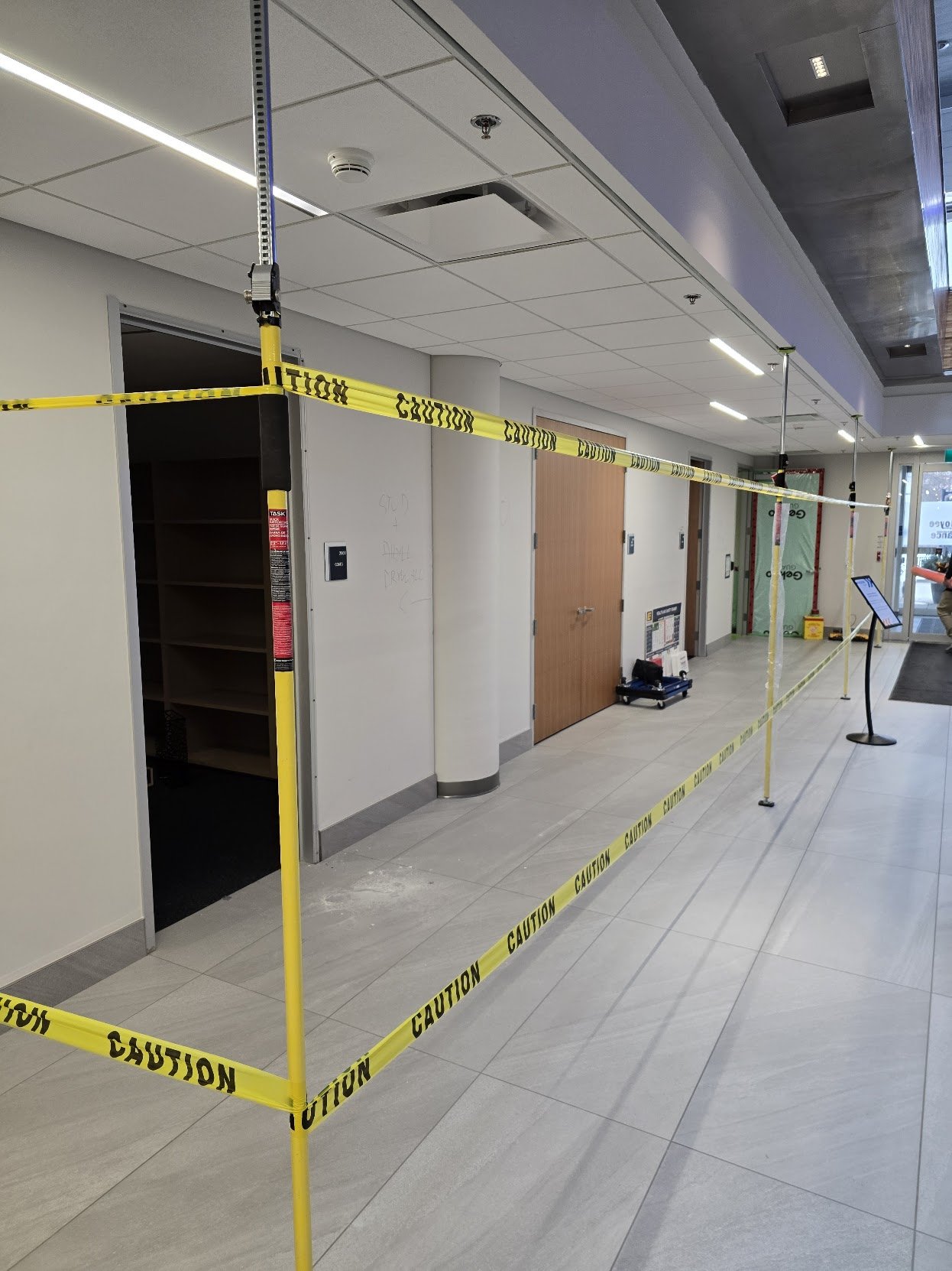 Yellow caution tape cordons off a section of the corridor inside a building, with open doors and a sign stand nearby.