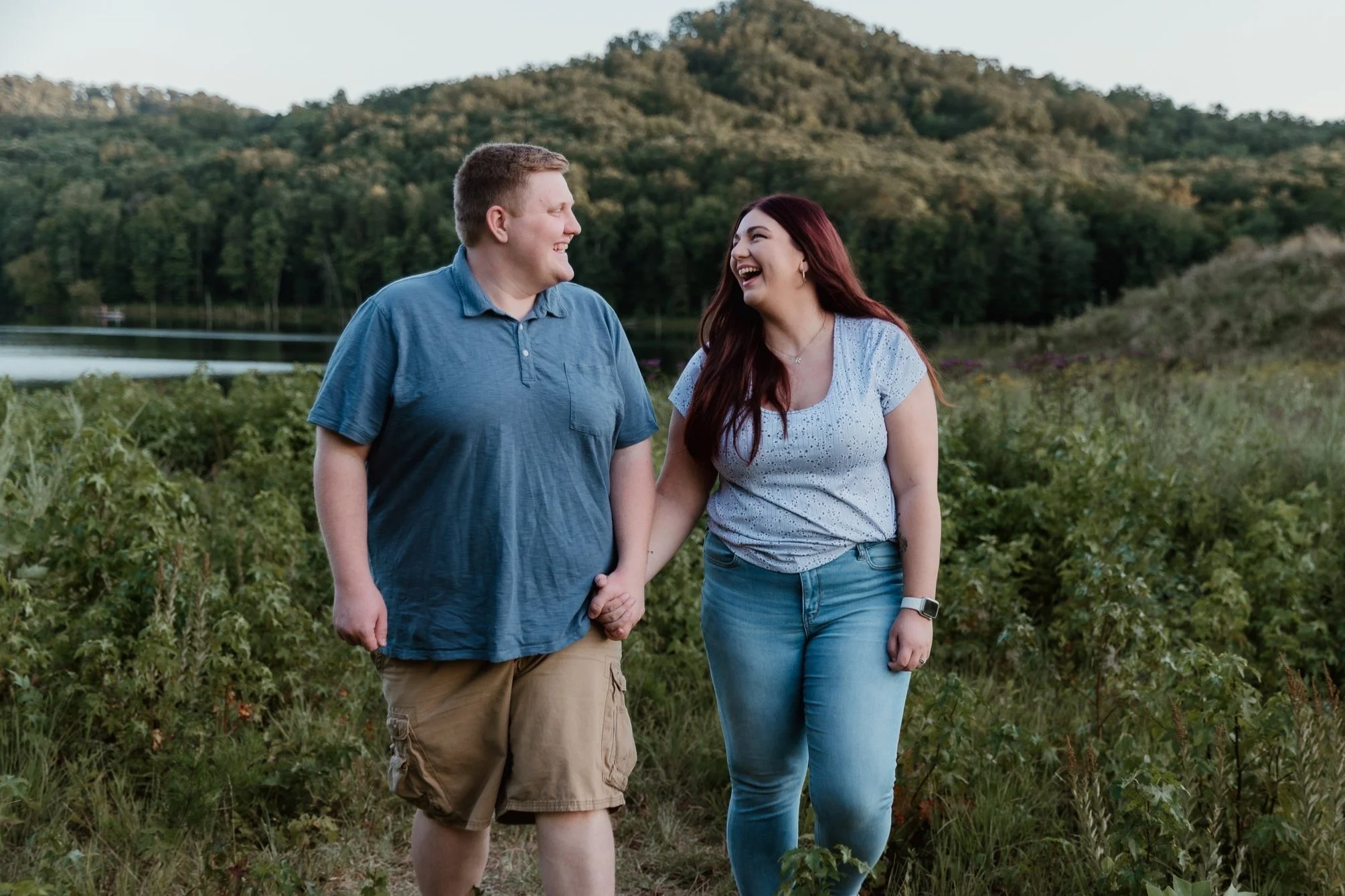 A smiling couple holding hands, walking outdoors in a natural setting with green bushes, trees, a lake, and a hill in the background.