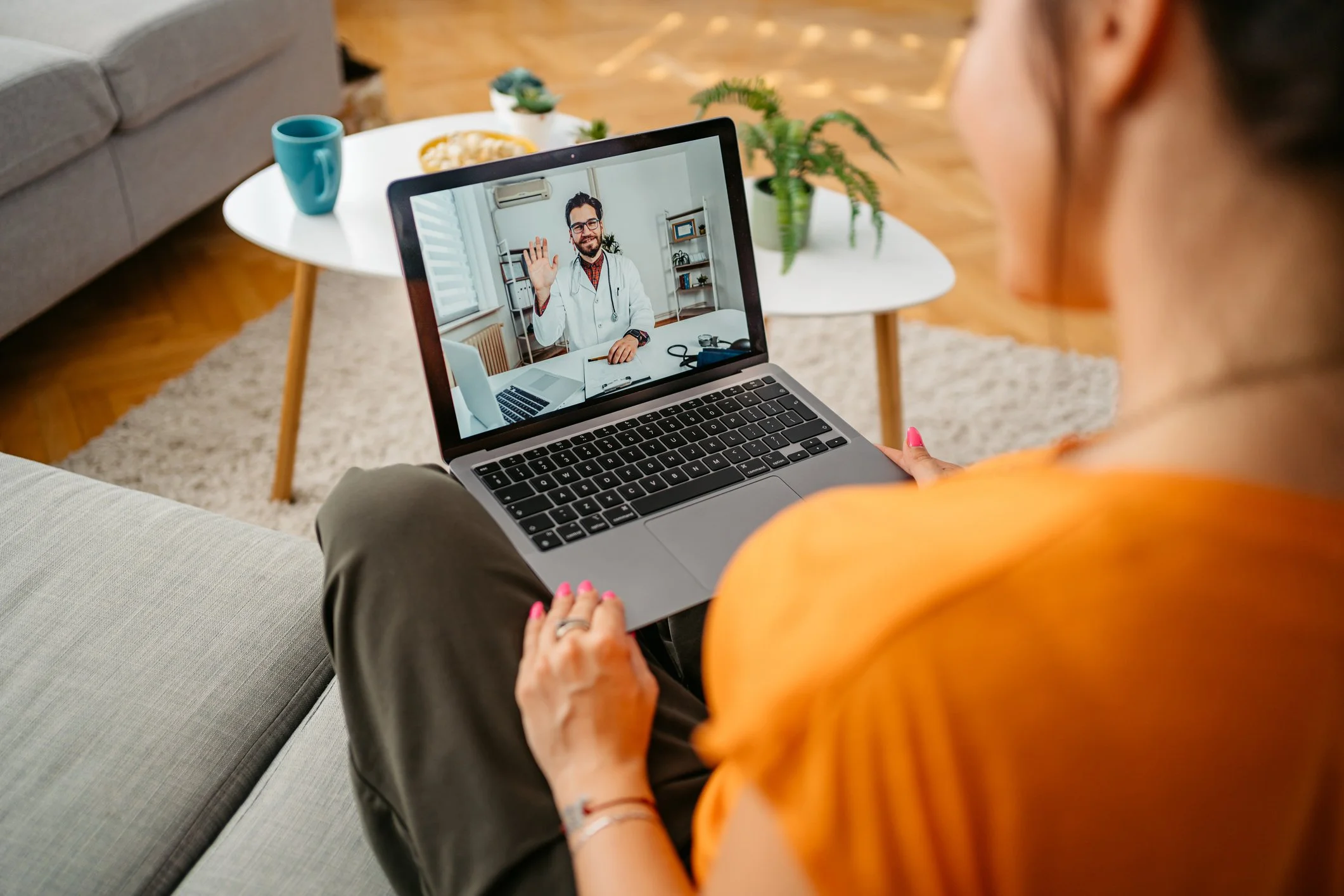 En kvinde ser på en laptop, hvor en mand i hvidt lægekit vinker til hende i et videokald.