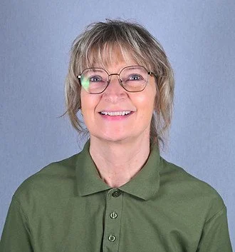 Et smilende ældre kvindemenneske med briller og kort blond hår, iført en grøn polo-trøje, foran en blå baggrund.