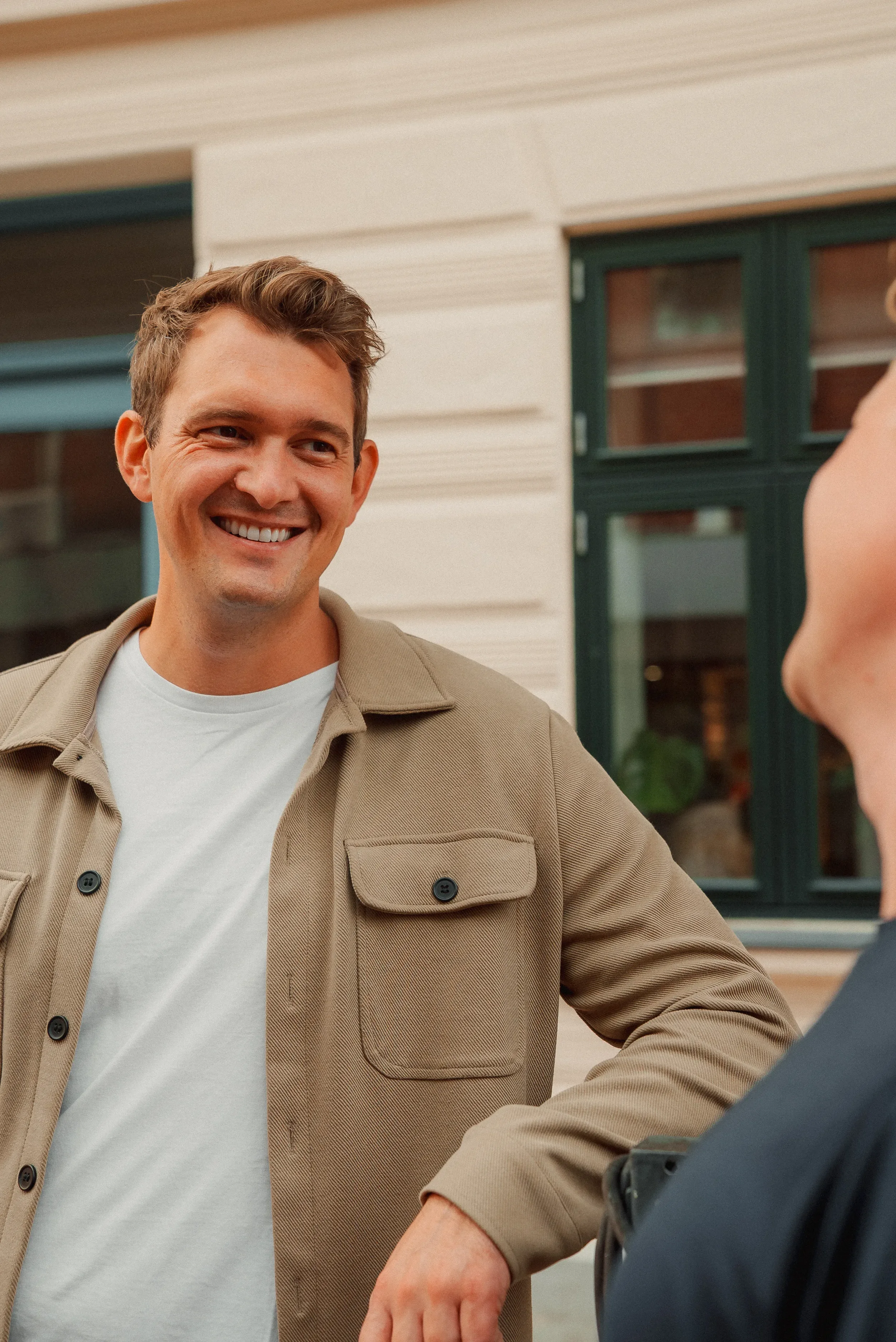 To personer snakker udenfor ved et hus med grønne vinduer. En person smiler.