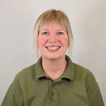 En kvinde med blond hår og grøn polo, smilende foran en hvid baggrund.