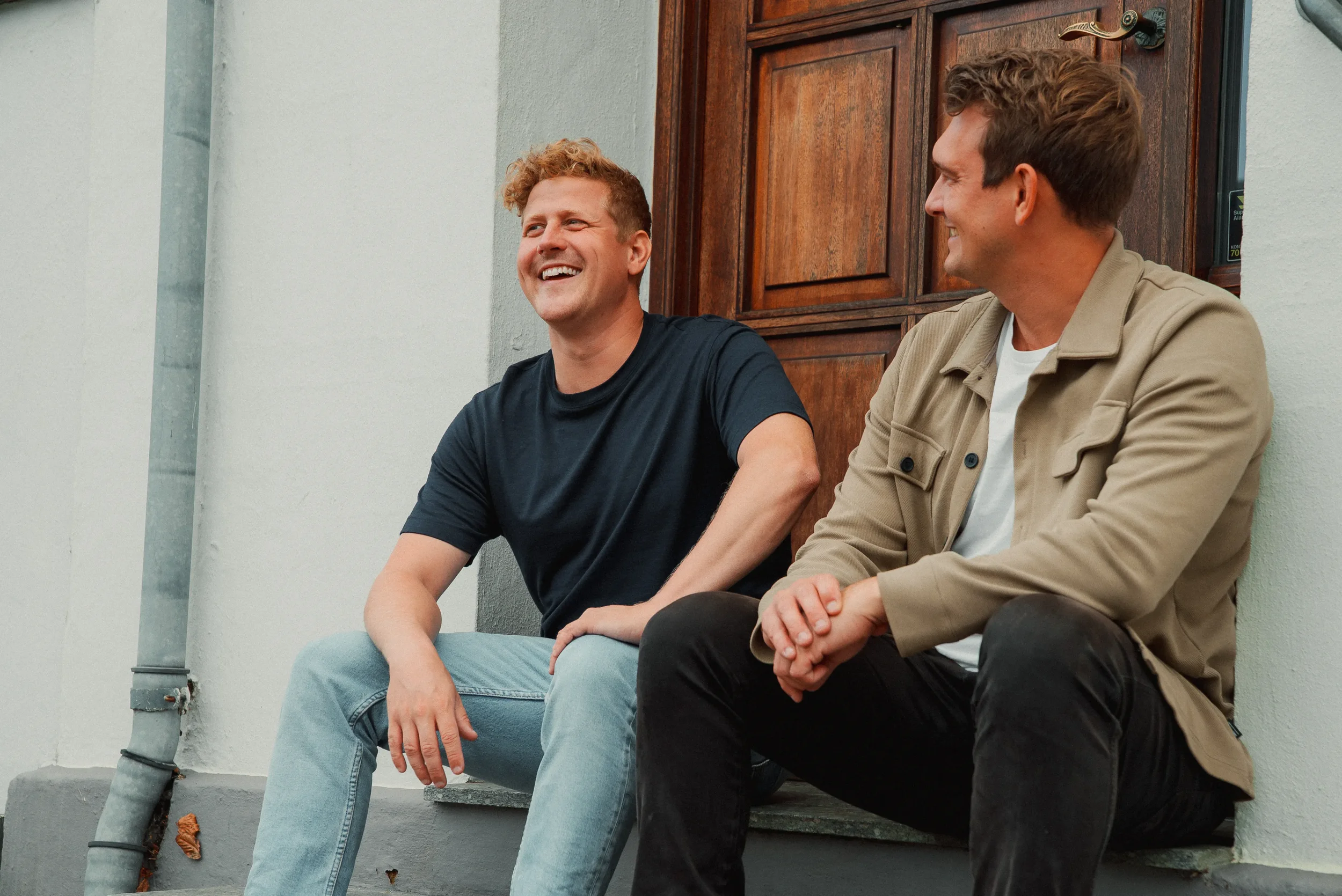To mænd sidder og snakker på trappen foran et hus med trædør og hvid facade, de smiler og har samtale.
