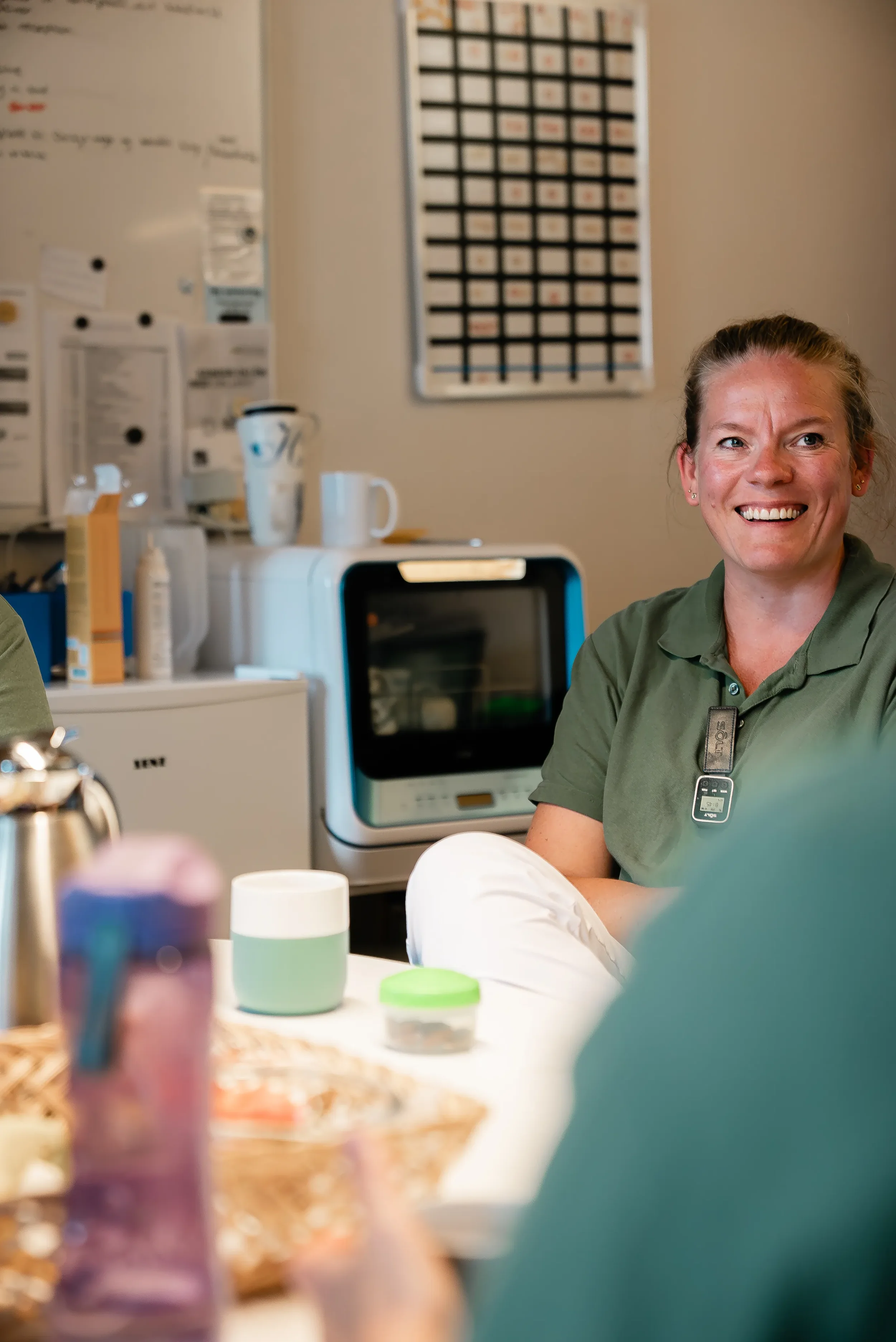 En smilende kvinde i en grøn arbejdsuniform sidder ved et bord i et kontormiljø med forskellige kopper, en plastboks, og en kaffemaskine i baggrunden.