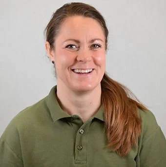 Et smilende portræt af en kvinde med brunt hår i en grøn polo mod en grå baggrund.