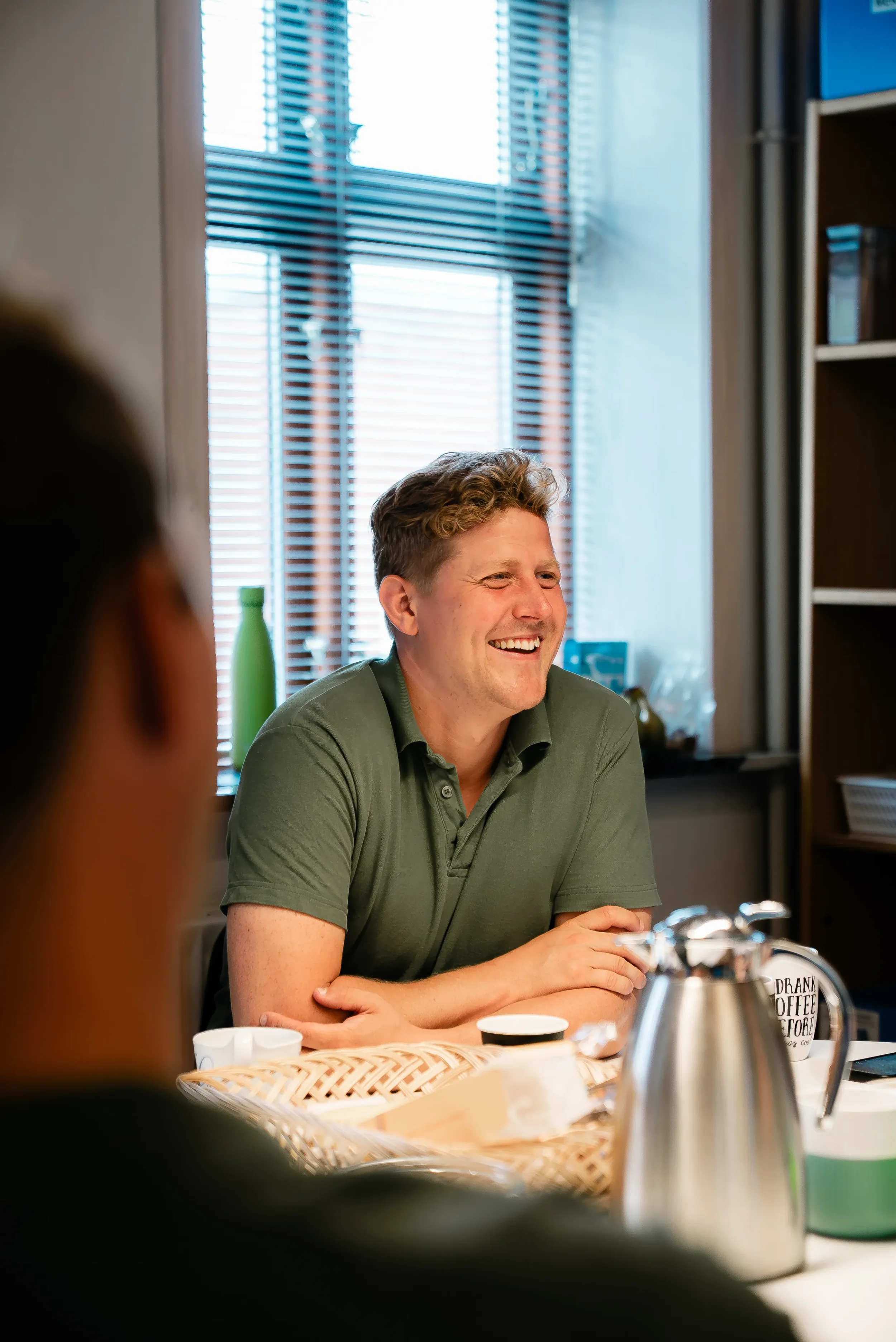 En mand sidder ved et bord og smiler, omgivet af kaffekander, kopper og snacks, indendørs med vinduer, der har vandrette blind.