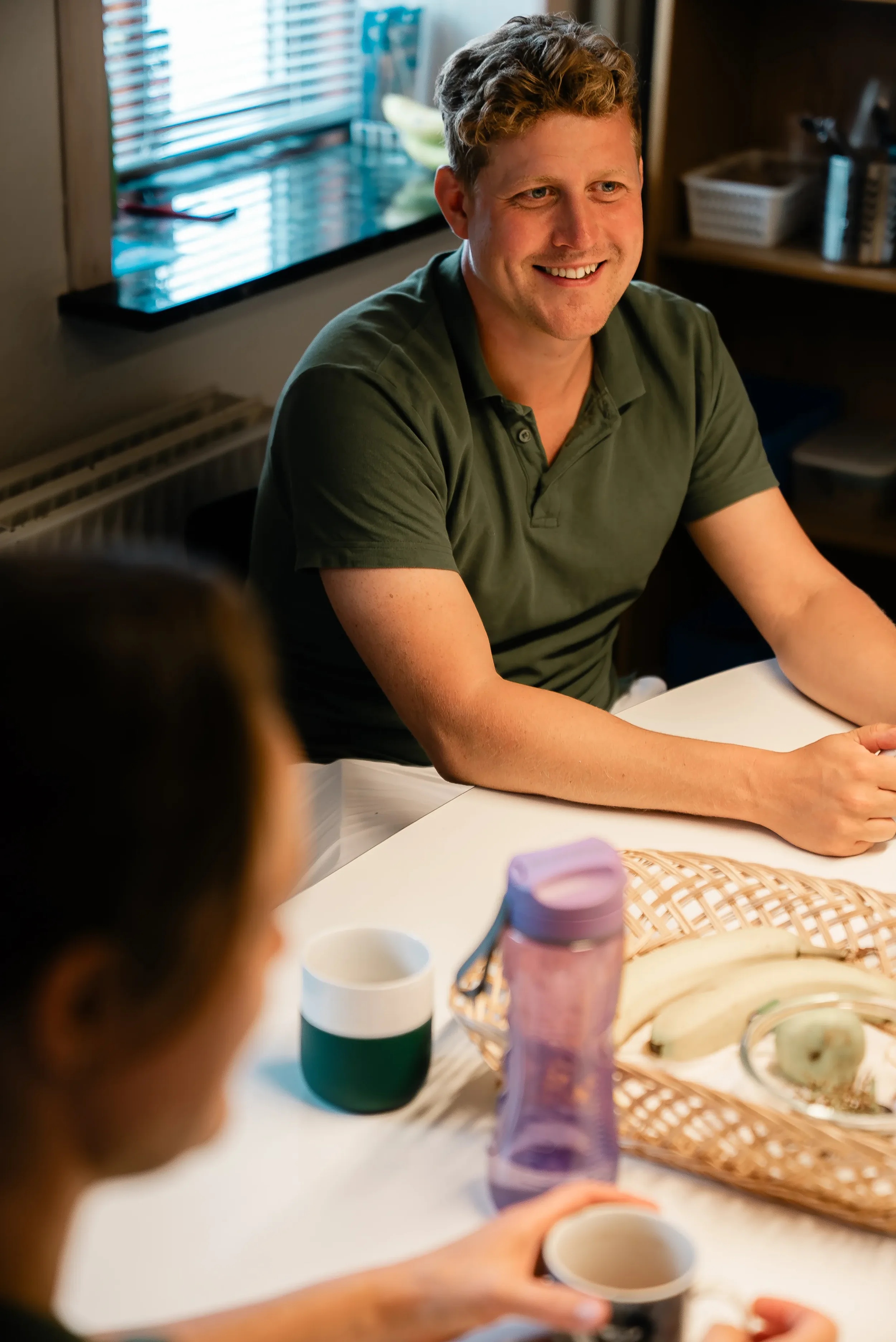 En mand med kort, bølget, blond hår smiler mens han sidder ved et spisebord med en kvinde, der holder en kop. Der er en skål med frugt og to krus på bordet, og solen skinner ind gennem vinduet bag.