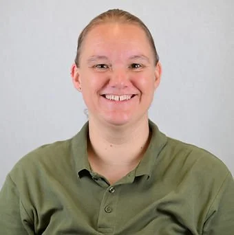 En smilende kvinde med lys hud og blond hår, iført en grøn polo mod lys grå baggrund.