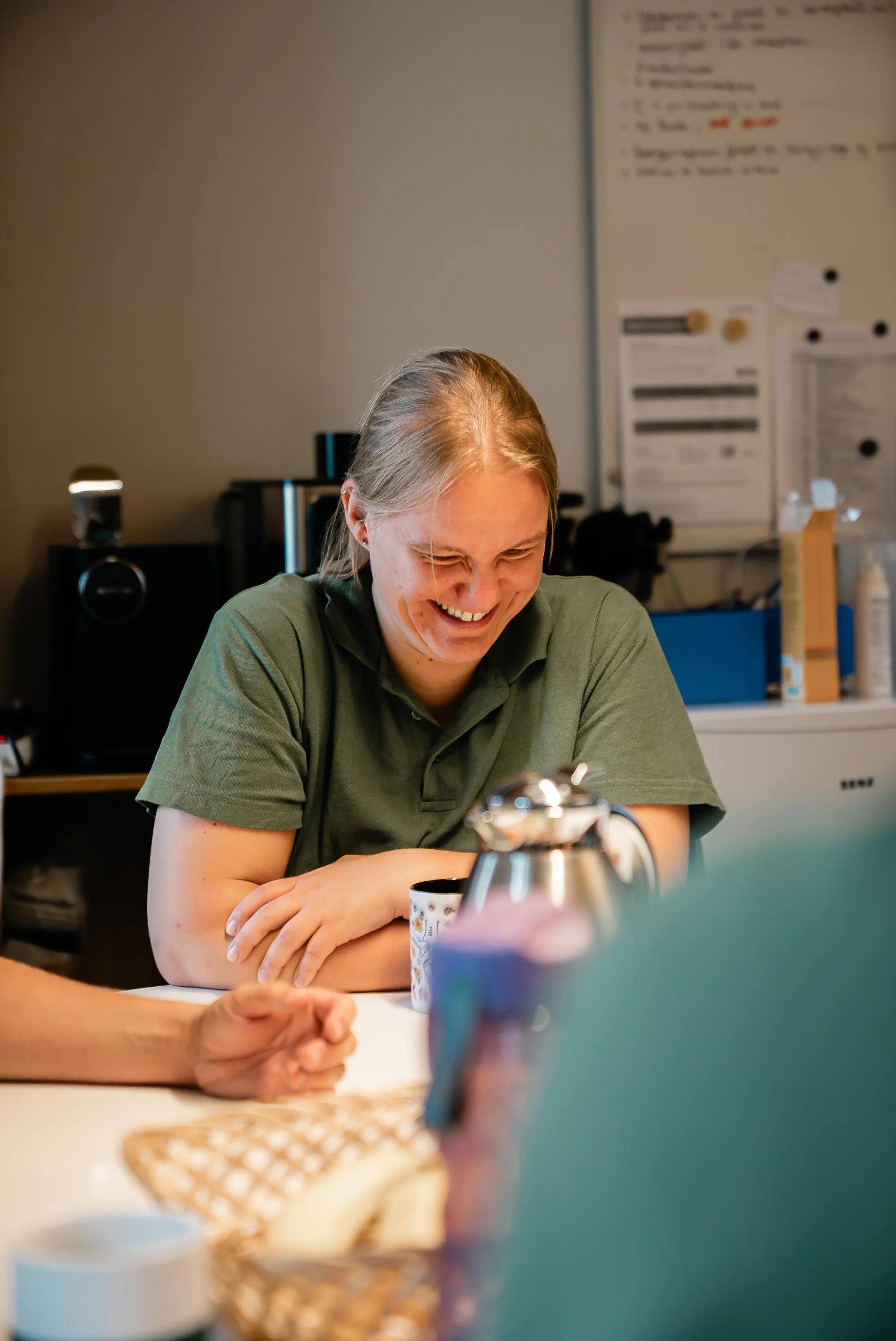En kvinde med langt blondt hår smiler og ler, mens hun sidder ved et bord med en kop, en tepotte og andre genstande. Bag hende er der en whiteboard med skrift og kontorudstyr.