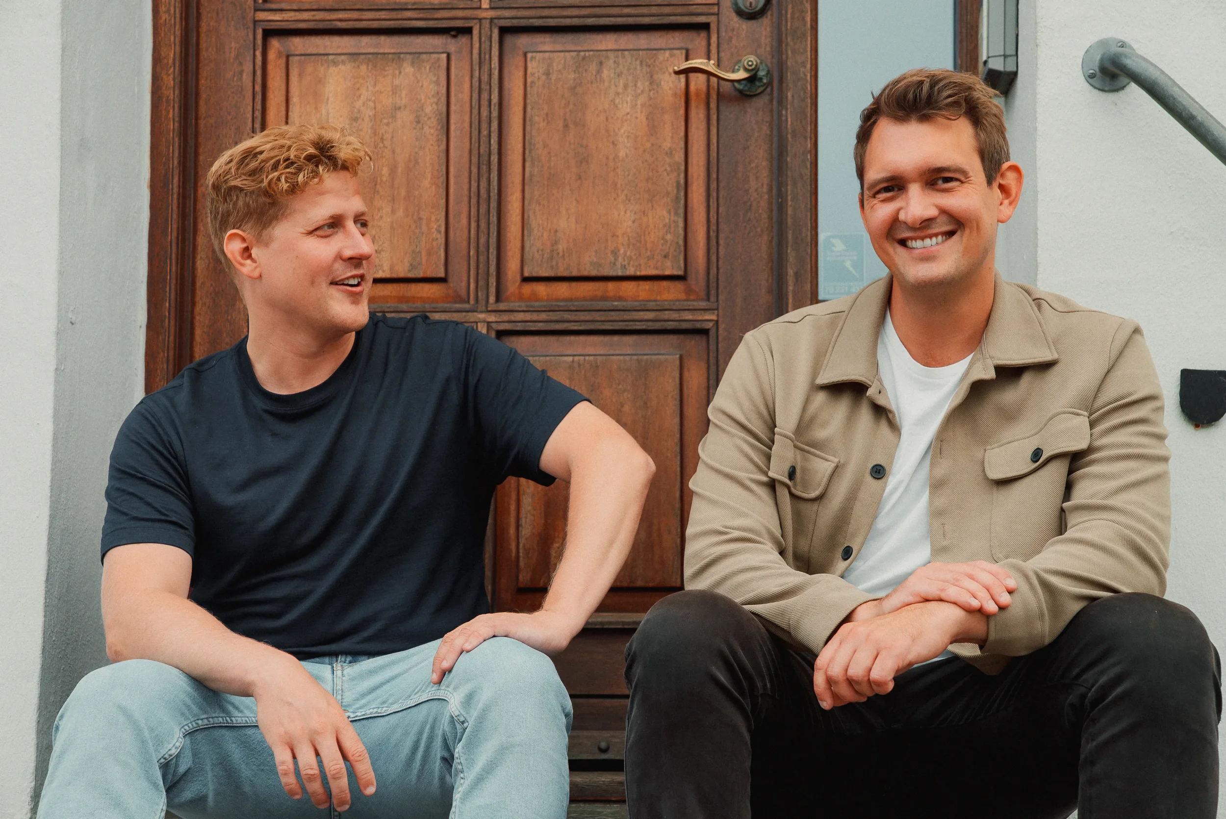 To unge mænd sidder på trappen foran en dør, de smiler og taler med hinanden.