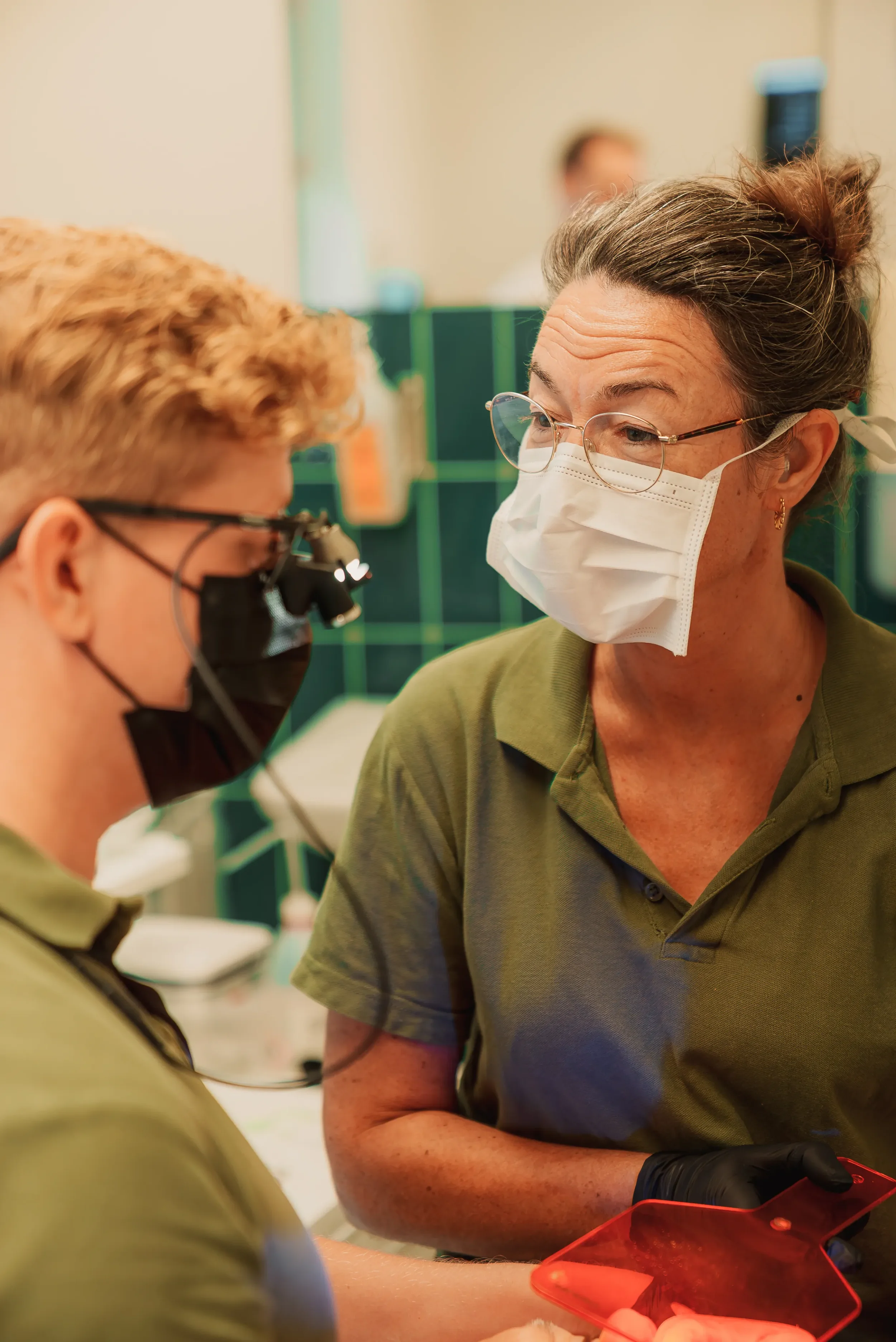 To kvinder, begge iført masker og briller, er i et medicinsk miljø. Den ene holder en rød enhed, mens de ser på noget sammen.