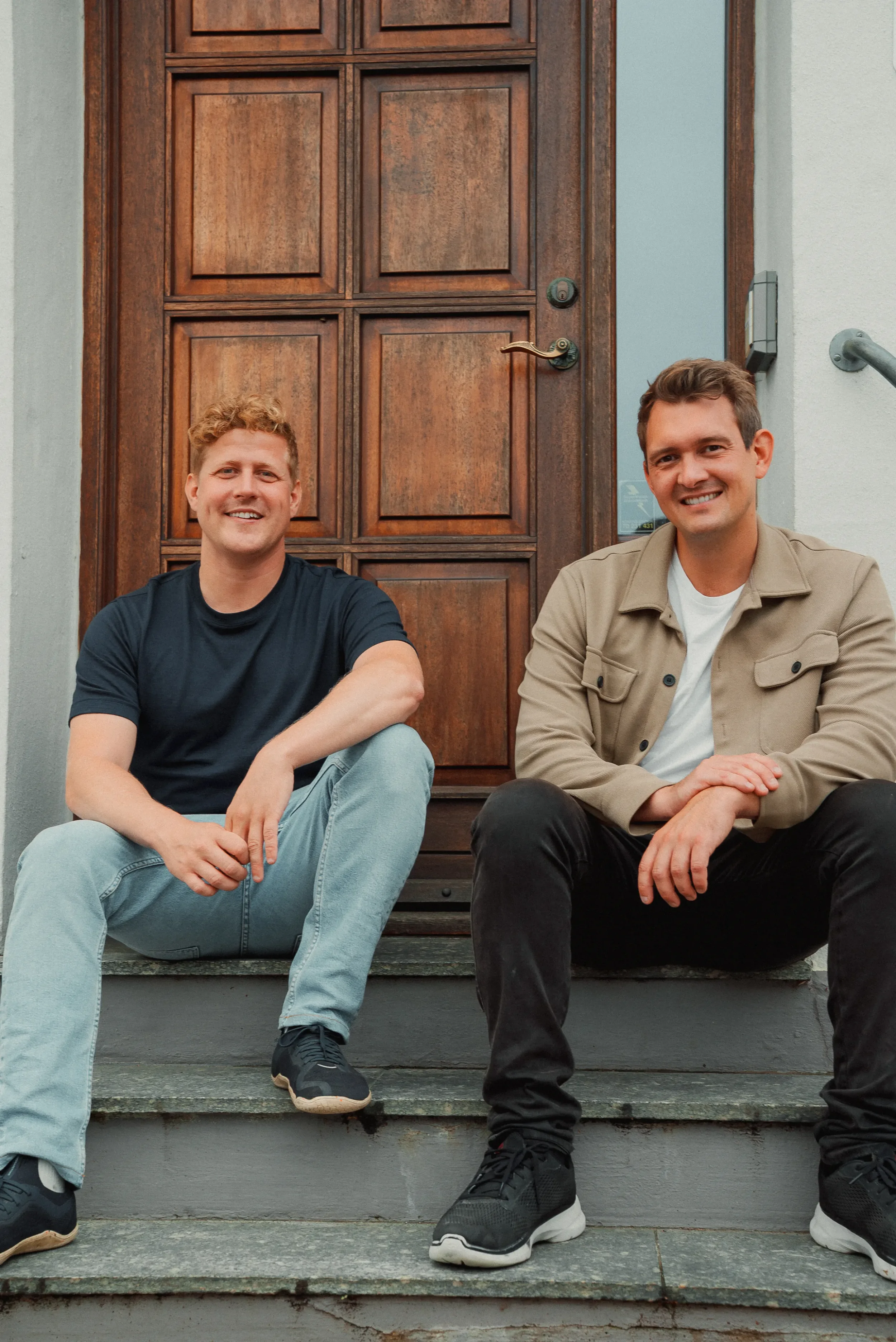 To unge mænd sidder på trappesten foran en trædør, smilende mod kameraet.