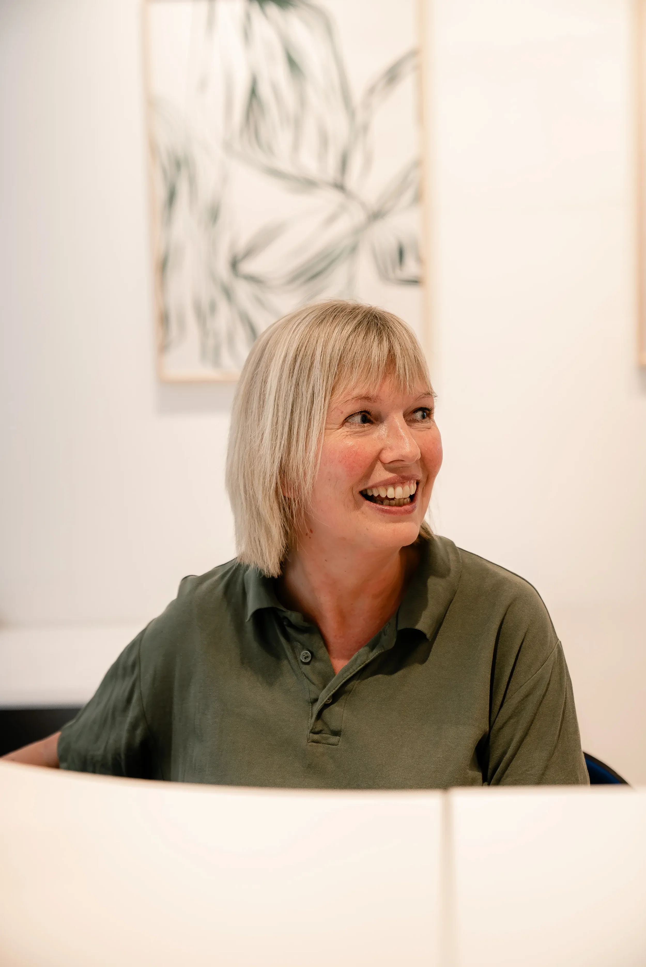 Kvinde med blond hår, smilende, i en grøn polo, siddende foran en hvidskærm, baggrund med abstrakt kunst på væggen.