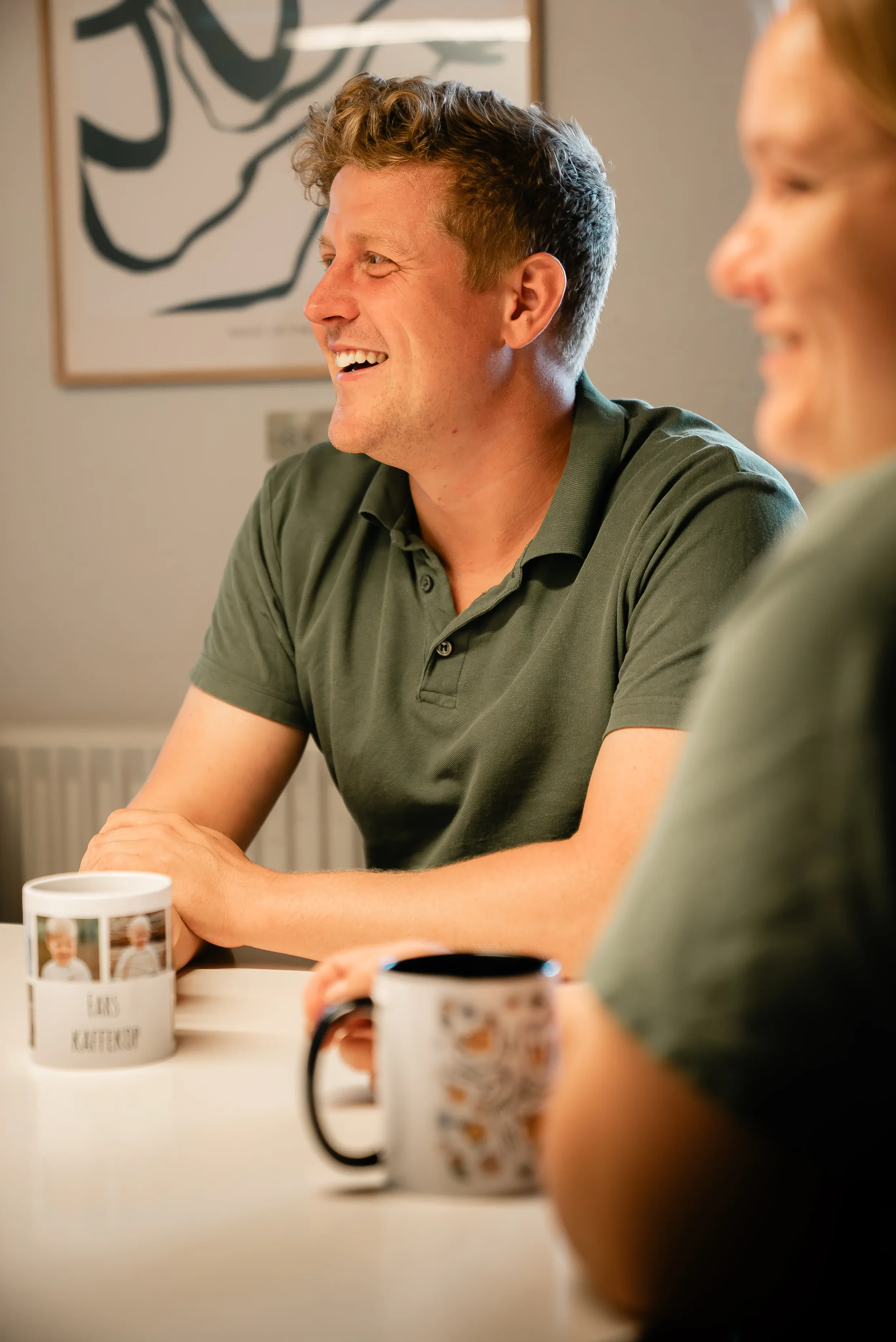 To personer sidder ved et bord, der er kommet kaffe i kopper foran dem, og de smiler og taler sammen.