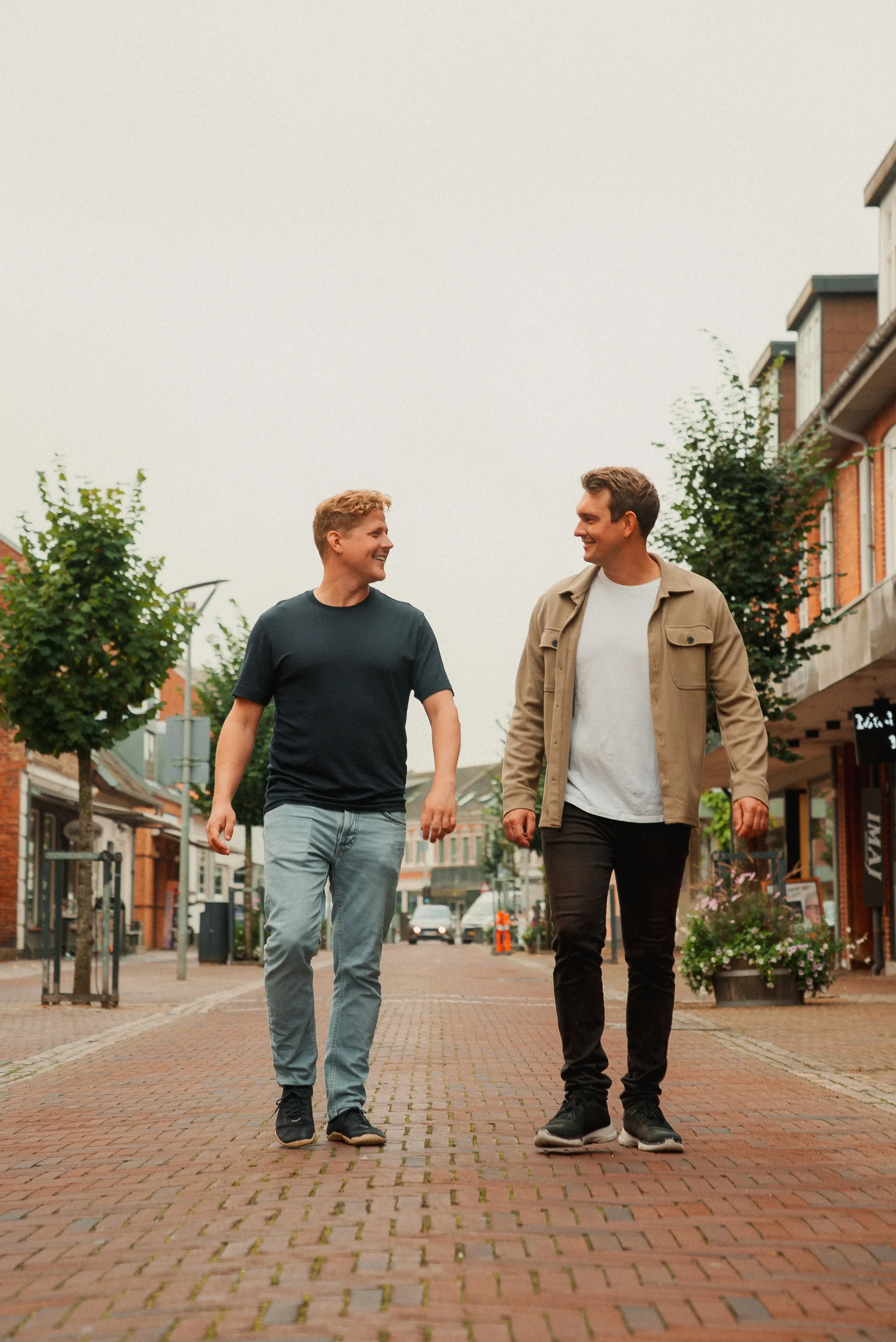 To mænd går sammen og smiler på en brostensbelagt gade i en by.