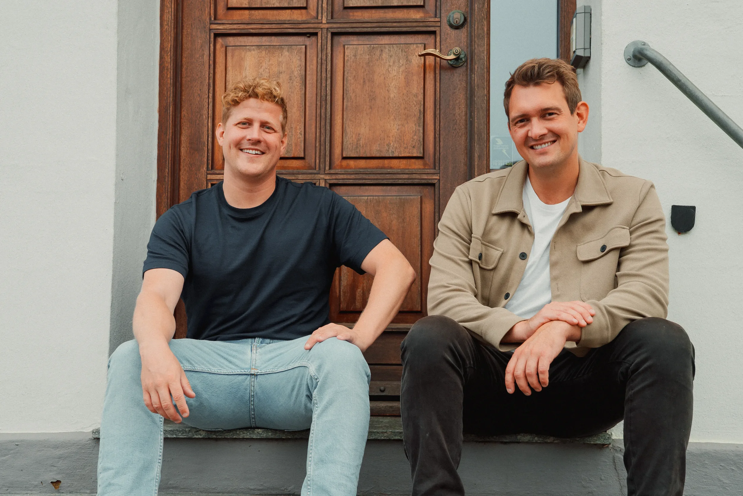 To mænd sidder på trappesten foran en trædør, smilende mod kameraet, ved tandlægehuset Bramming