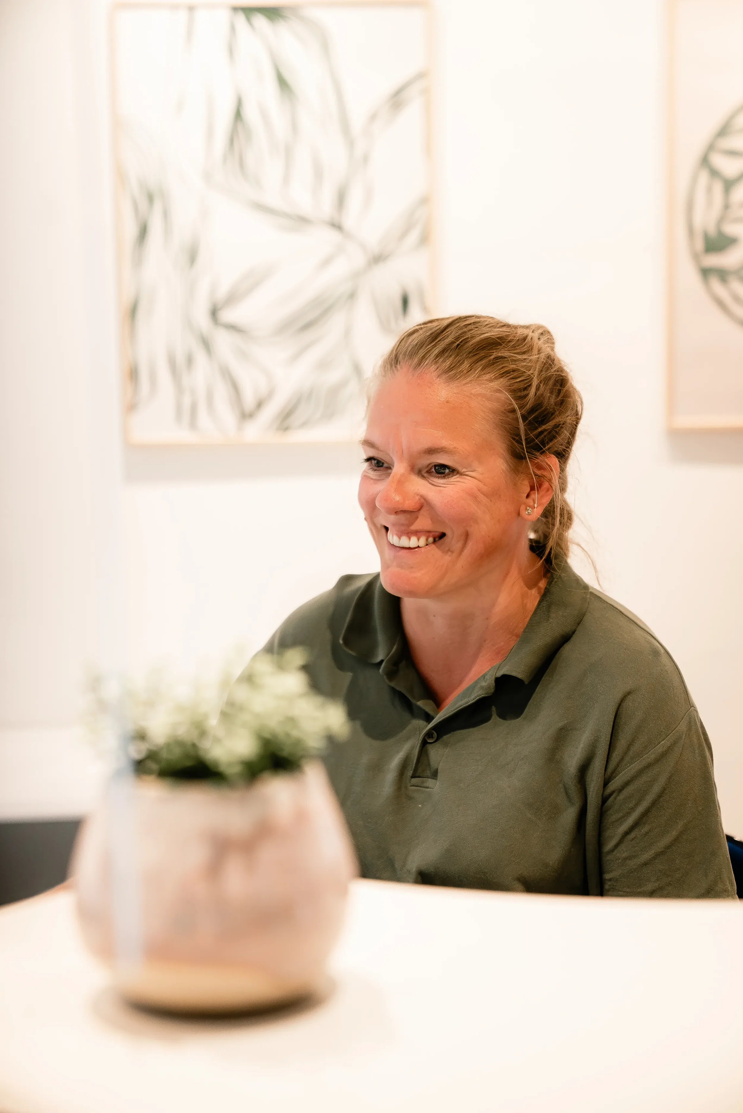 En smilende kvinde med lyst hår i en grøn polo, sidder ved et bord med en blomsterdekoration, med abstrakte grønne kunstværker på væggen bag hende.