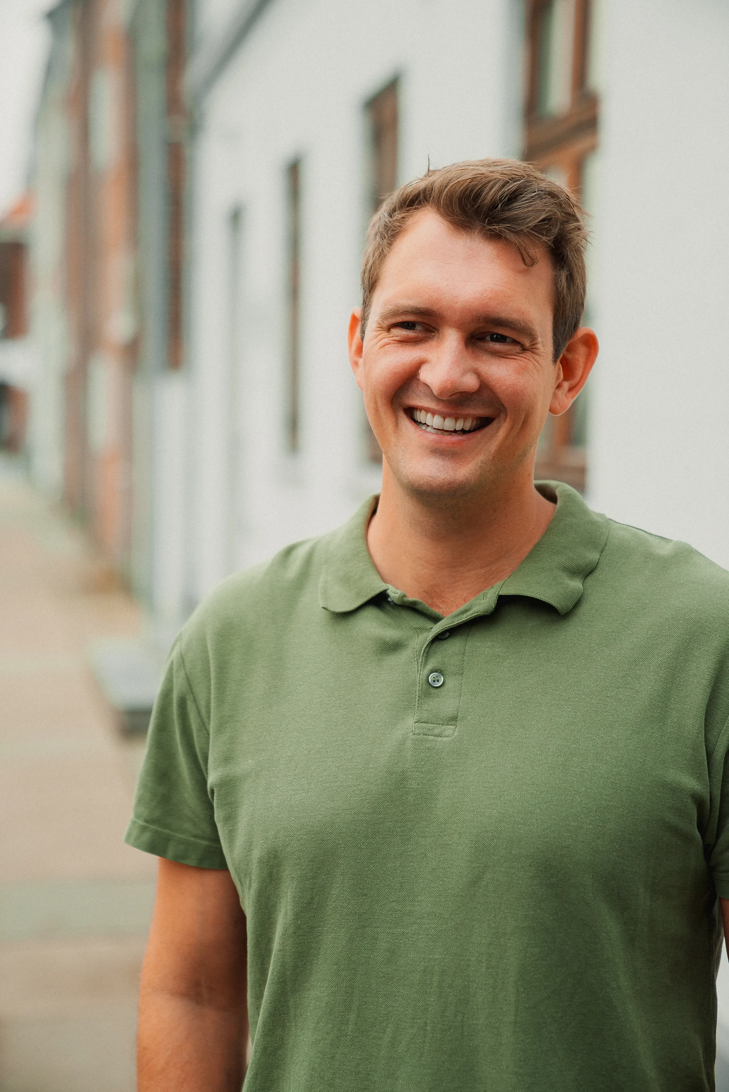 Ung mand med kort brunt hår i en grøn polo shirt smiler udendørs.