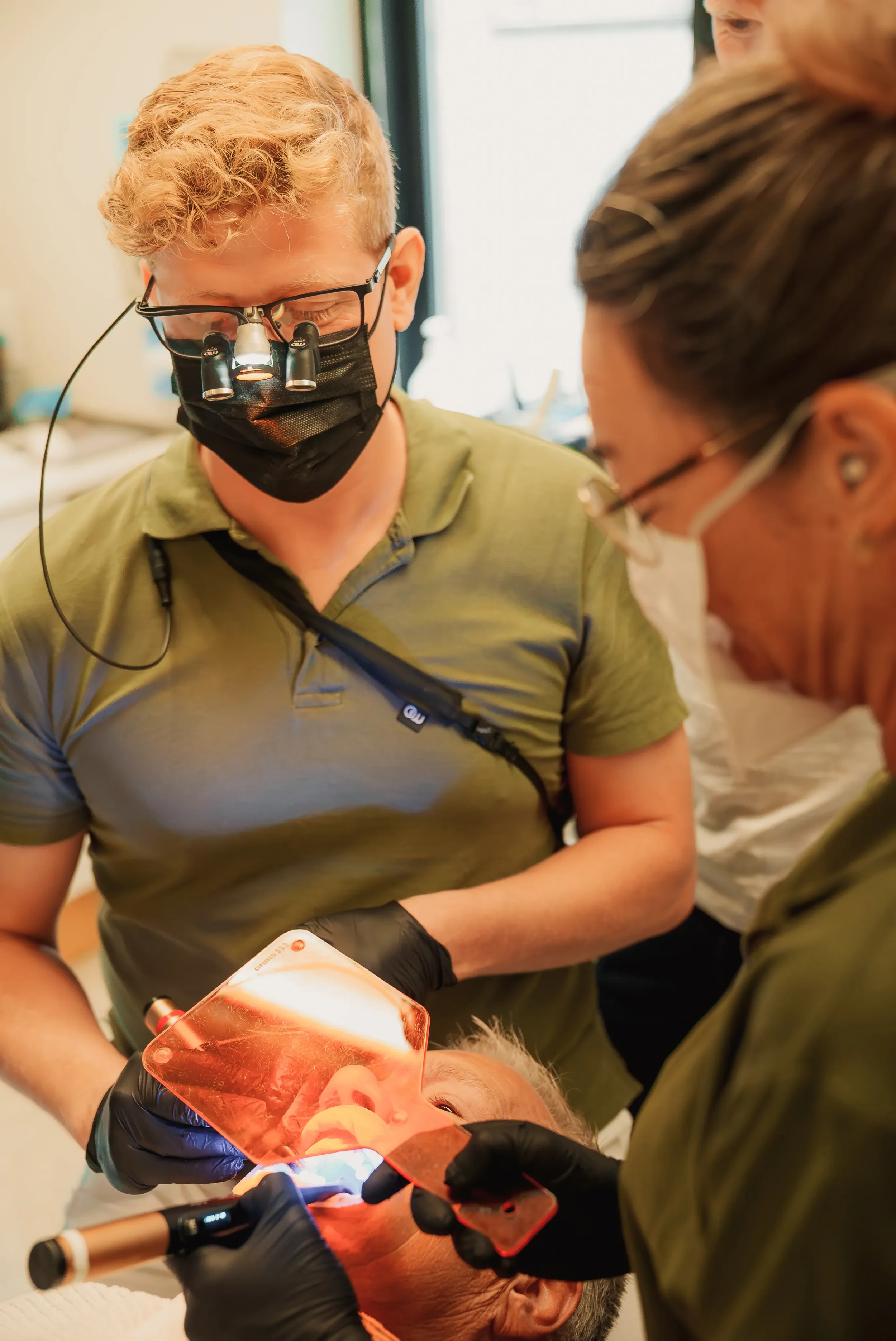 To læger i grønne kitler og masker arbejder med en patient i en medicinsk indretning, med forskningsudstyr og en stor lyskilde.