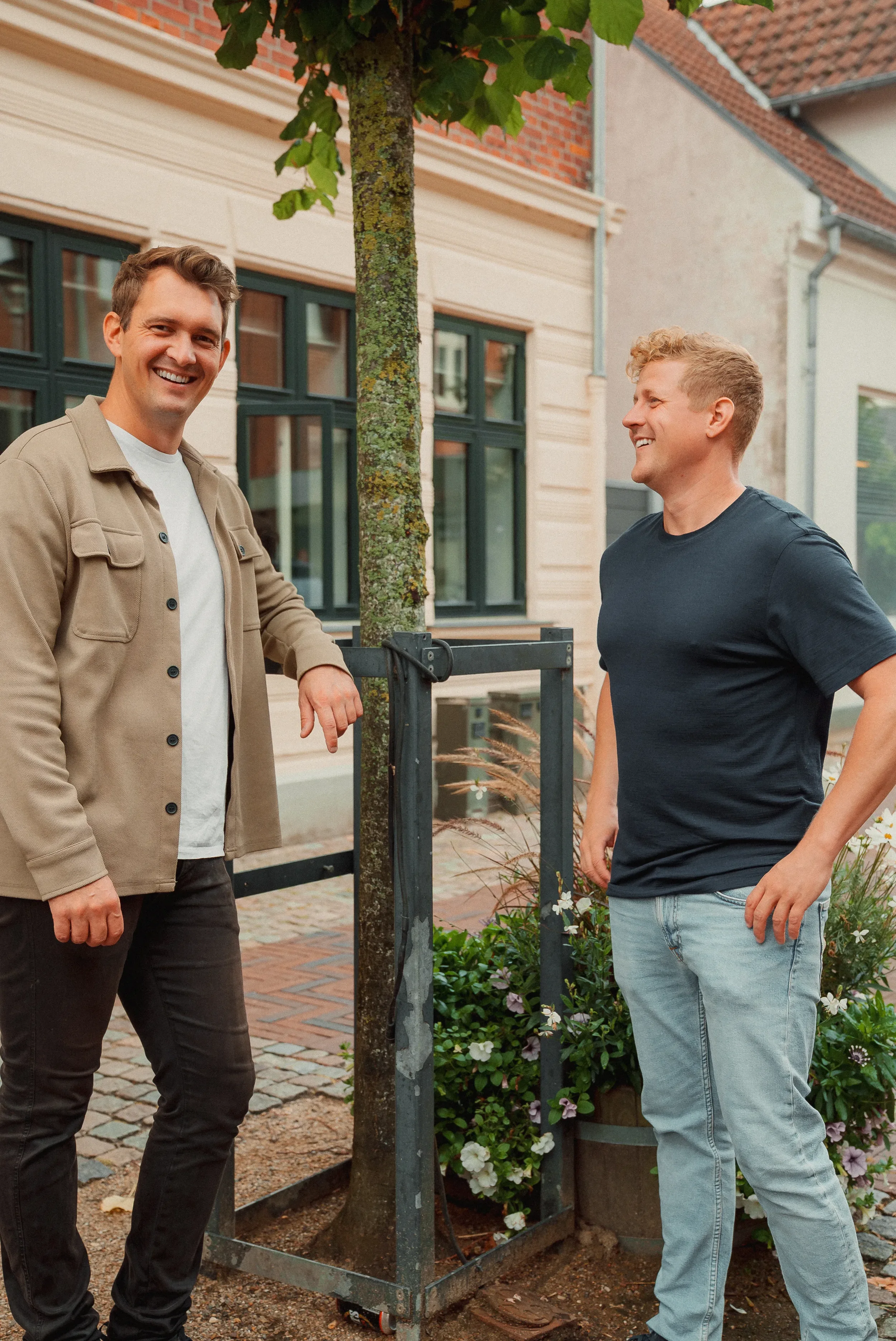 To mænd står ved en nyplantet træ i byen. Den ene mand har brunt hår og en beige jakke, den anden har blond hår og en sort T-shirt. De smiler og snakker.