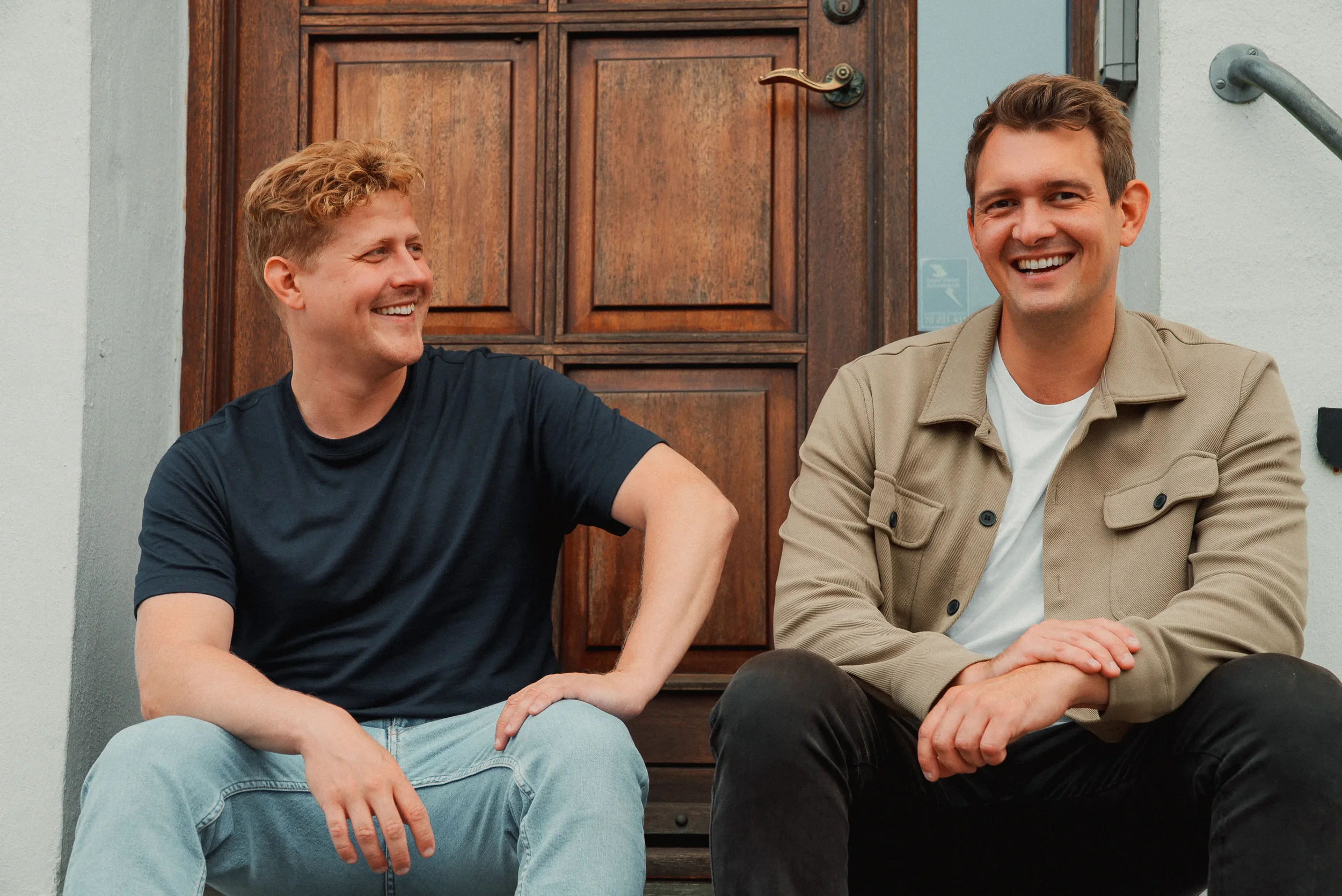 To mænd sidder på trappen foran en dør, de smiler og taler med hinanden.