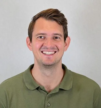 Et portrætbillede af en mand med brunt, kort hår, der smiler, iført en grøn polo i en neutral baggrund.