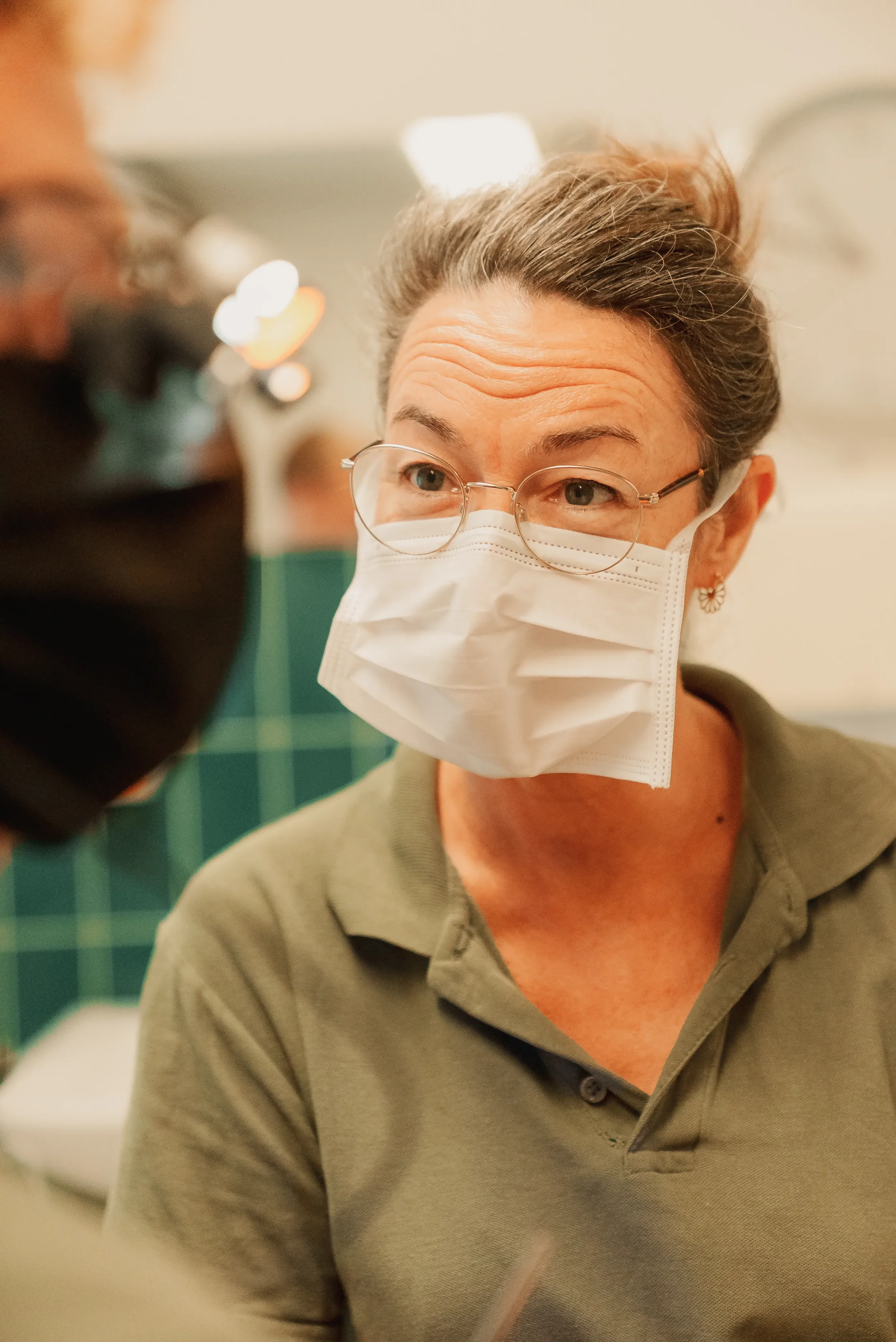 En kvinde med grå hår, briller, og øreringe, der taler med en person iført sort maske, begge bærer ansigtsmasker, i et indendørs miljø.