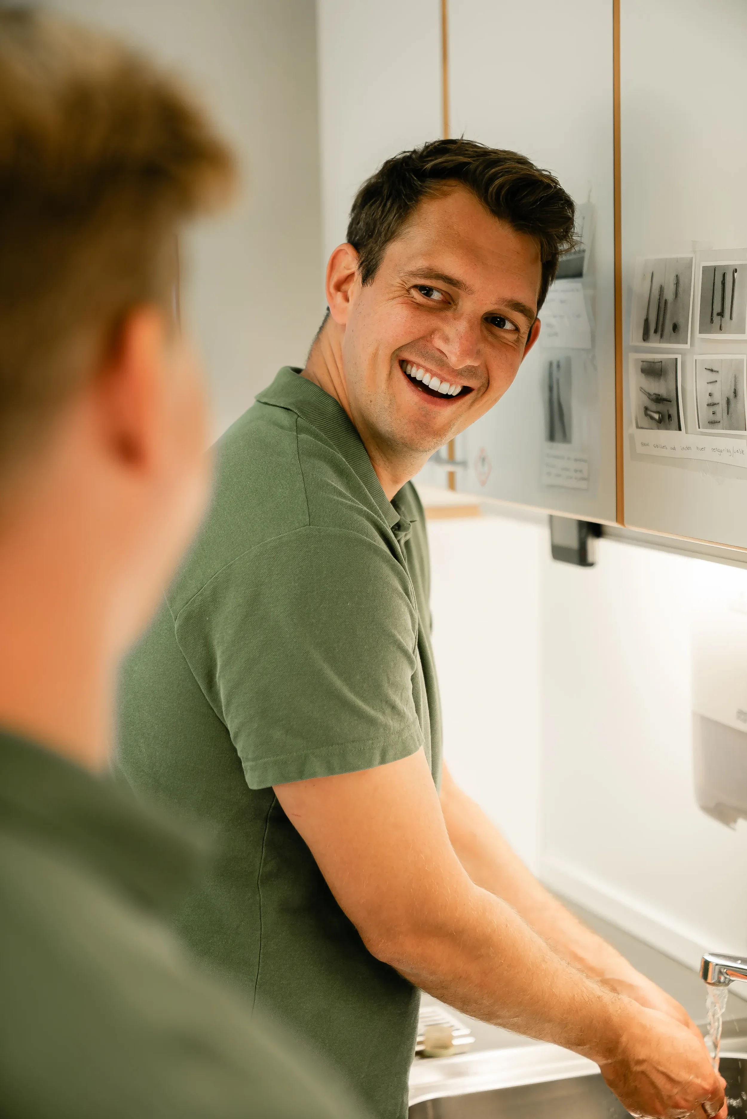 To mænd vasker hænder under en vask, den ene vender sig mod kamera og smiler.