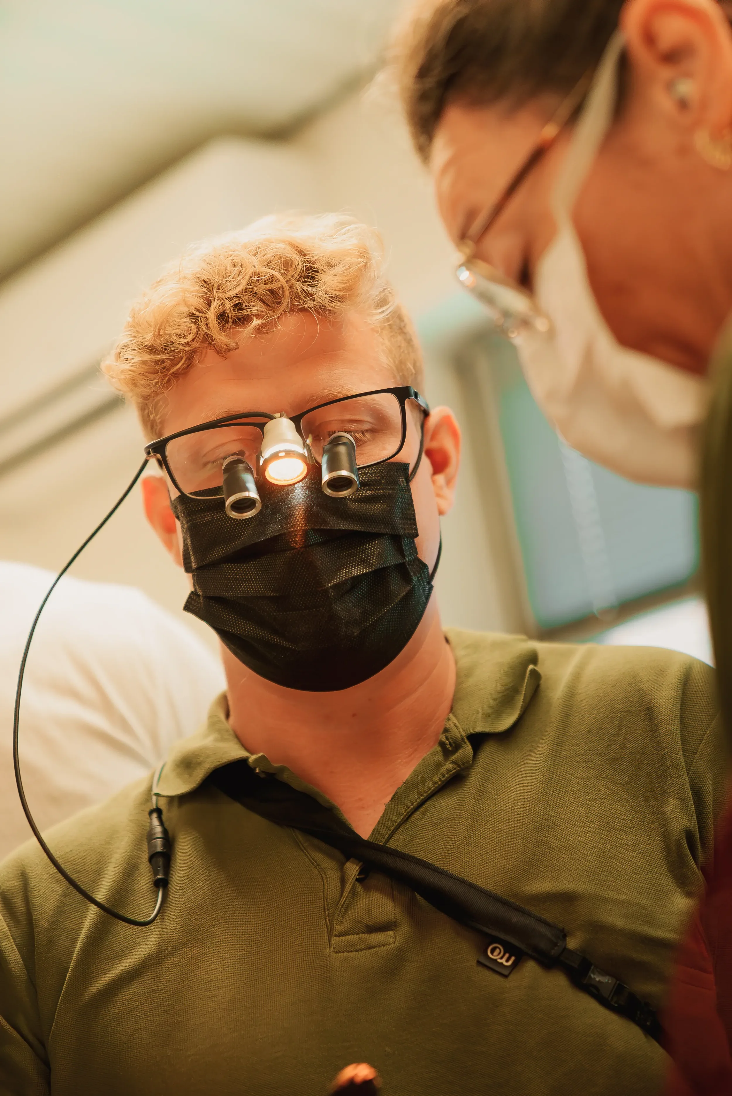 En mand iført grøn polo-shirt og ansigtmaske med forstørrelsesglas, der undersøger noget med professionel lup i et medicinsk eller dentalt sammenhæng, overvåget af en kvinde.